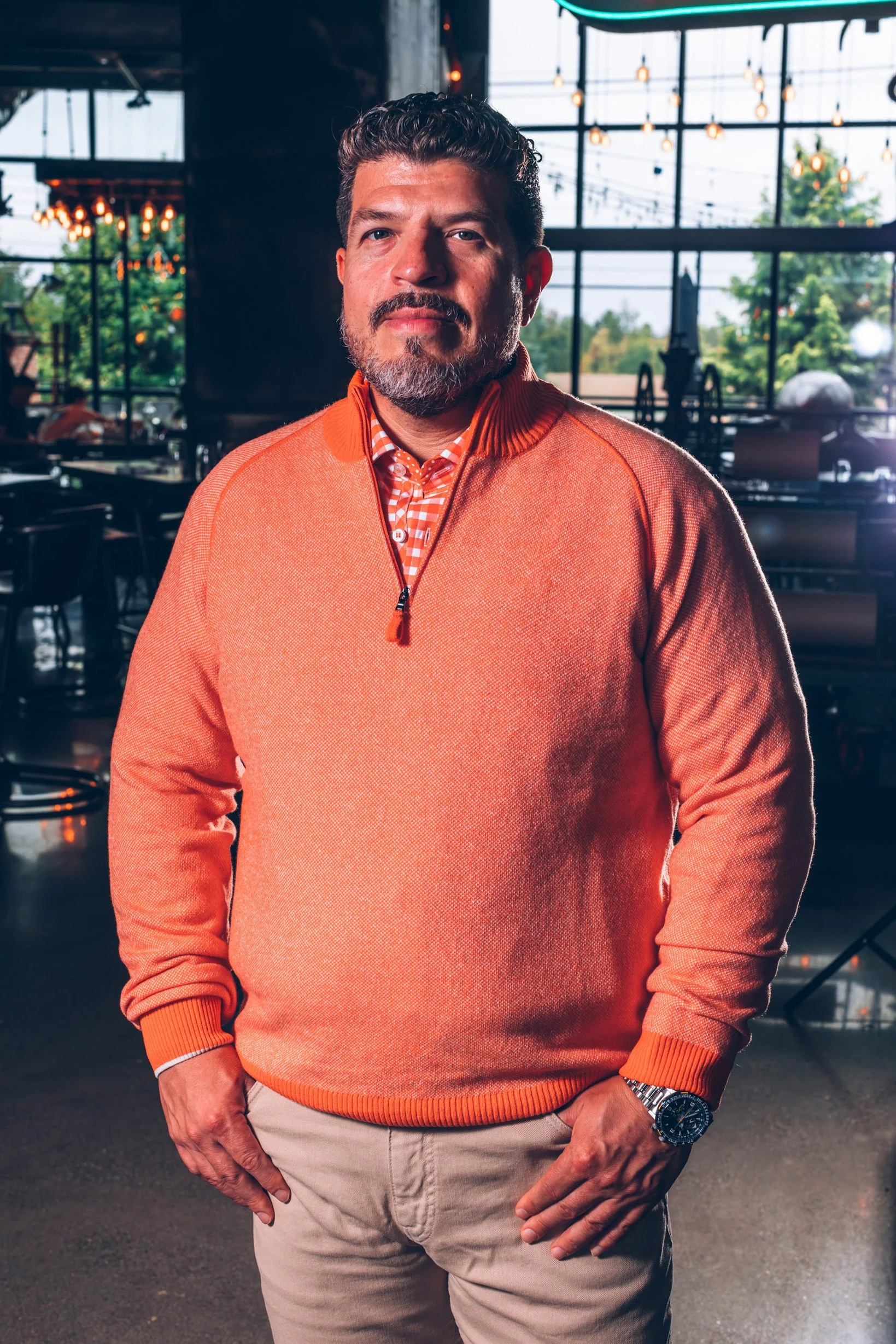 A man wearing an orange sweater, checked shirt, and beige pants standing in a restaurant or cafe with large windows, hanging lights, and a view of trees outside.