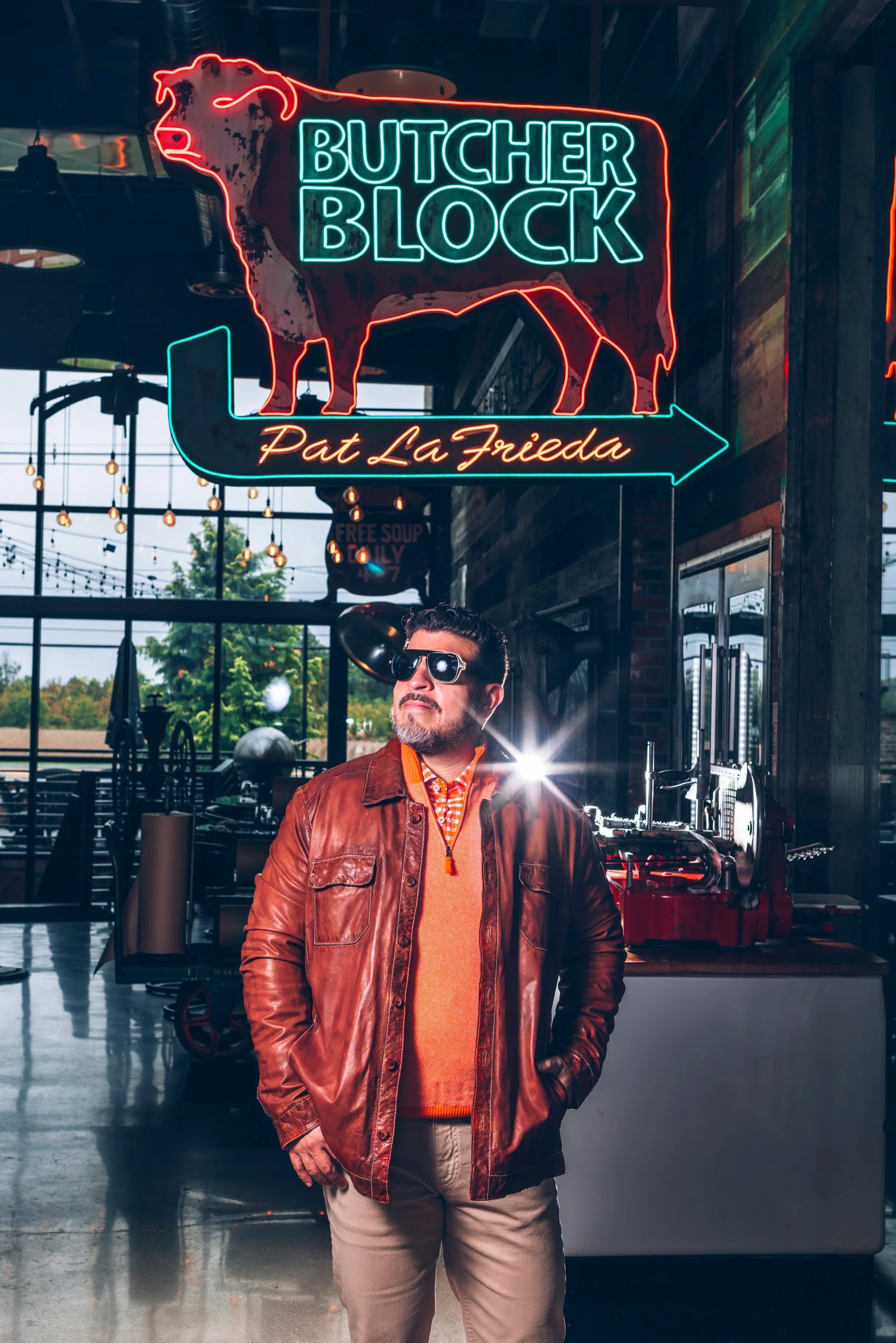A man with sunglasses and a leather jacket standing inside a restaurant or store with neon signs, including a cow outline and the words "Butcher Block" and "Pat LaFrieda".