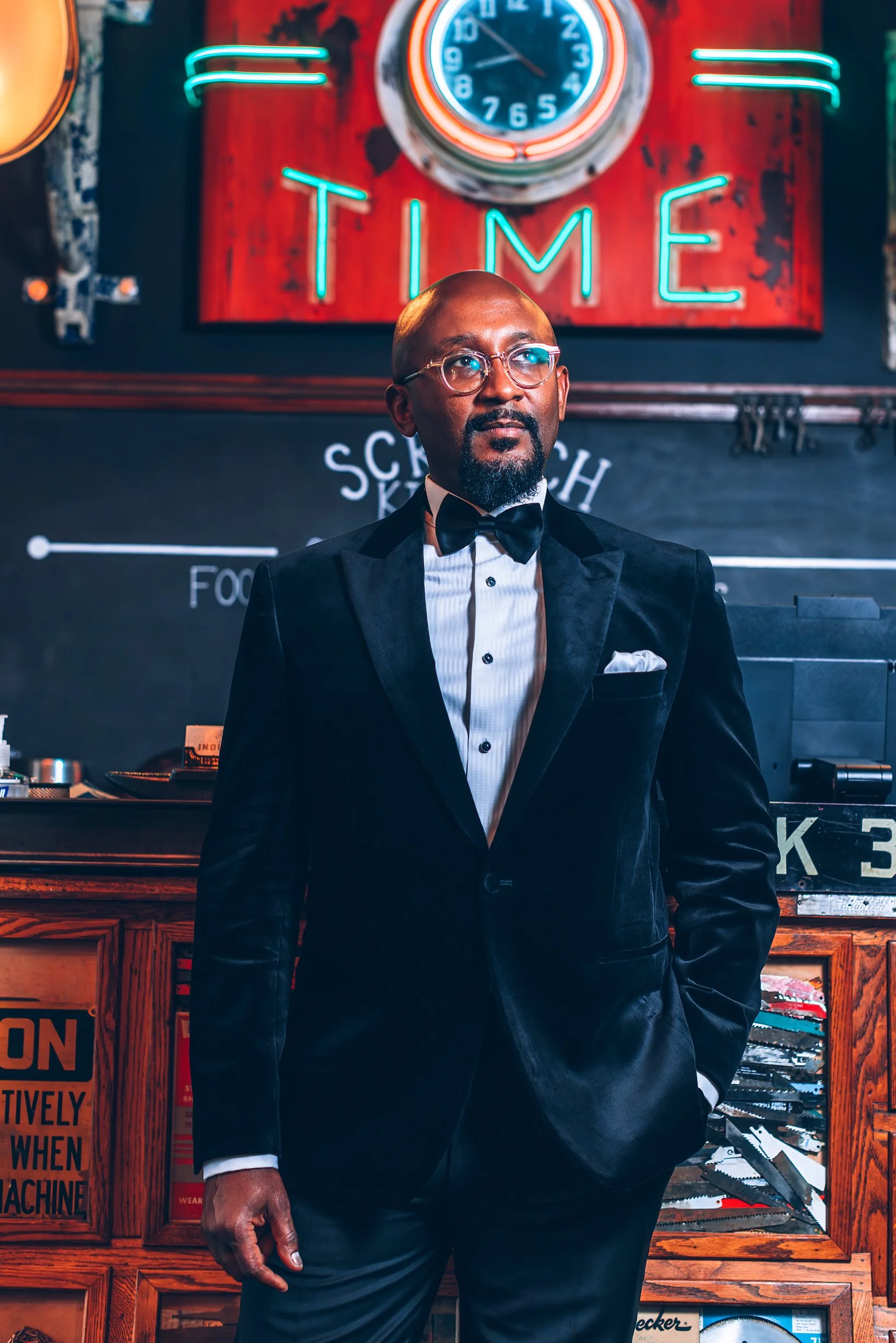 A man in a black tuxedo and bow tie standing in front of a chalkboard with the word 'TIME' in neon lights. The background features a clock and a wooden counter.