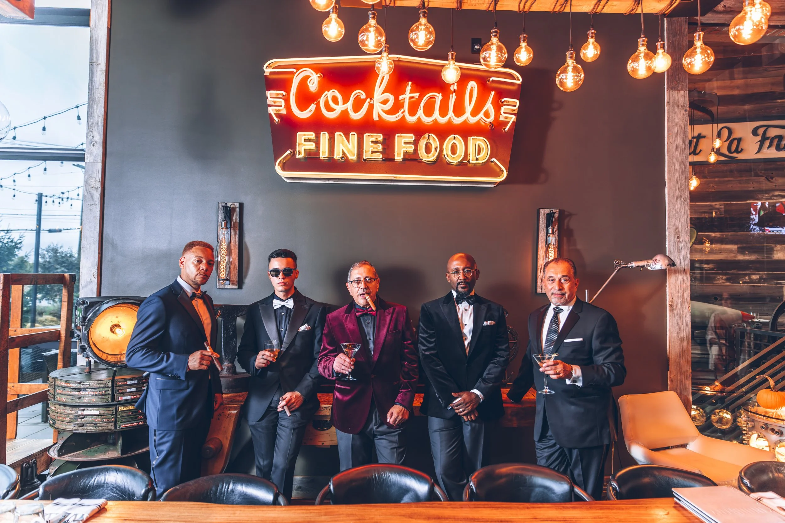 Five men in formal suits posing with cocktails in front of a neon sign that reads 'Cocktails' and 'Fine Food' in a stylish restaurant or bar with warm lighting and wooden decor.