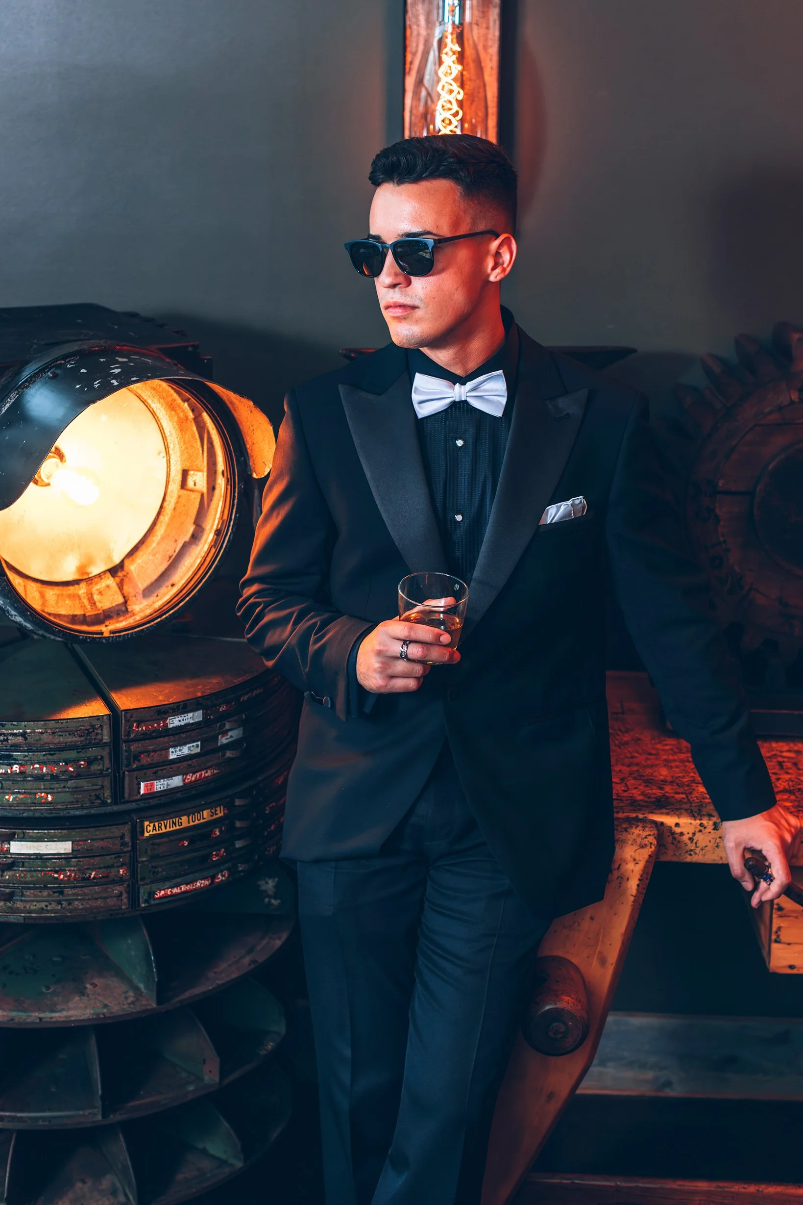 Man in tuxedo with sunglasses holding a glass, leaning on a vintage industrial machine in a dimly lit room.