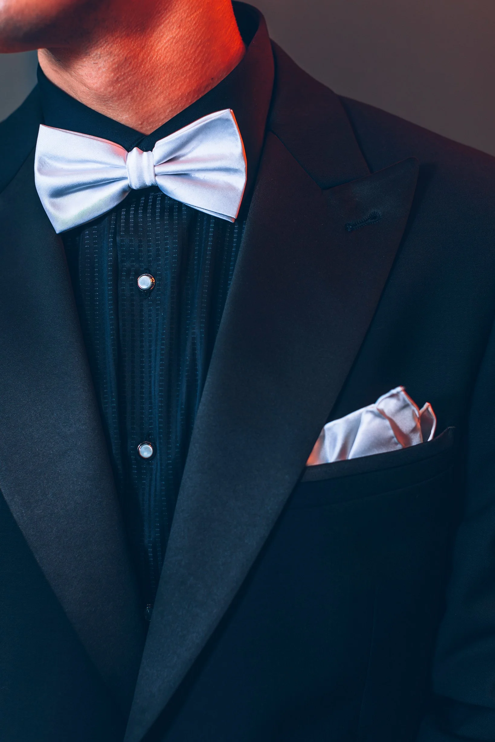 Close-up of a man wearing a black tuxedo with a white satin bow tie and matching pocket square, against a neutral background.