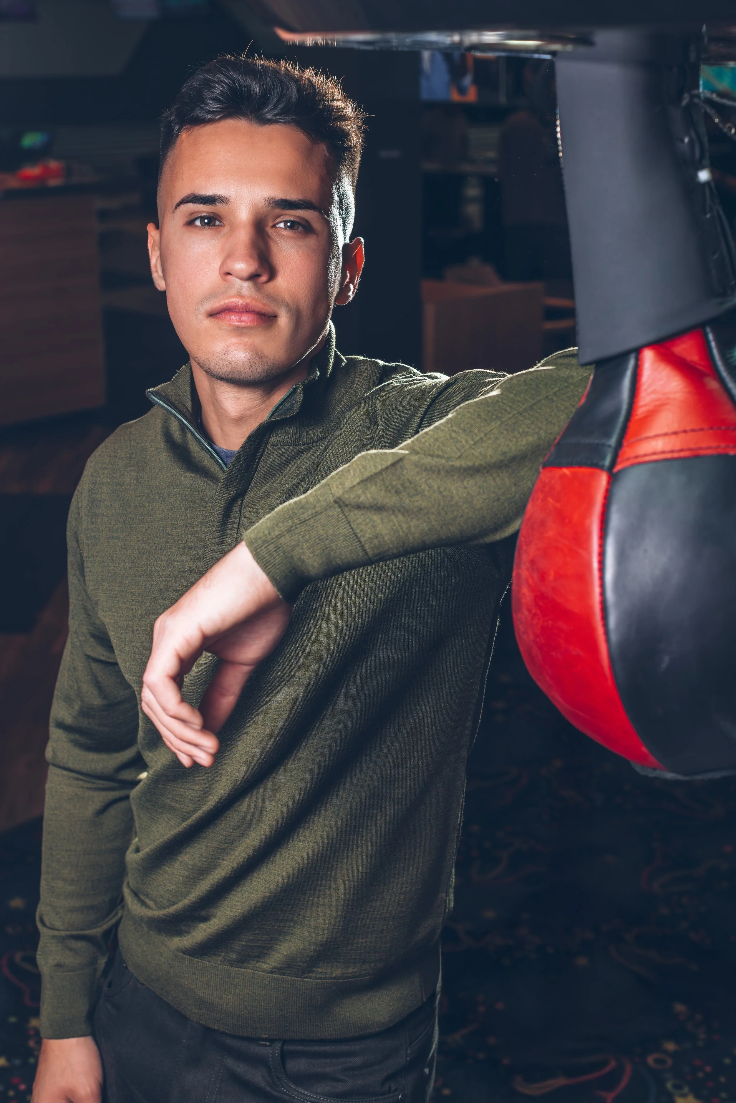 Young man in green sweater resting his arm on a punching bag in a gym.