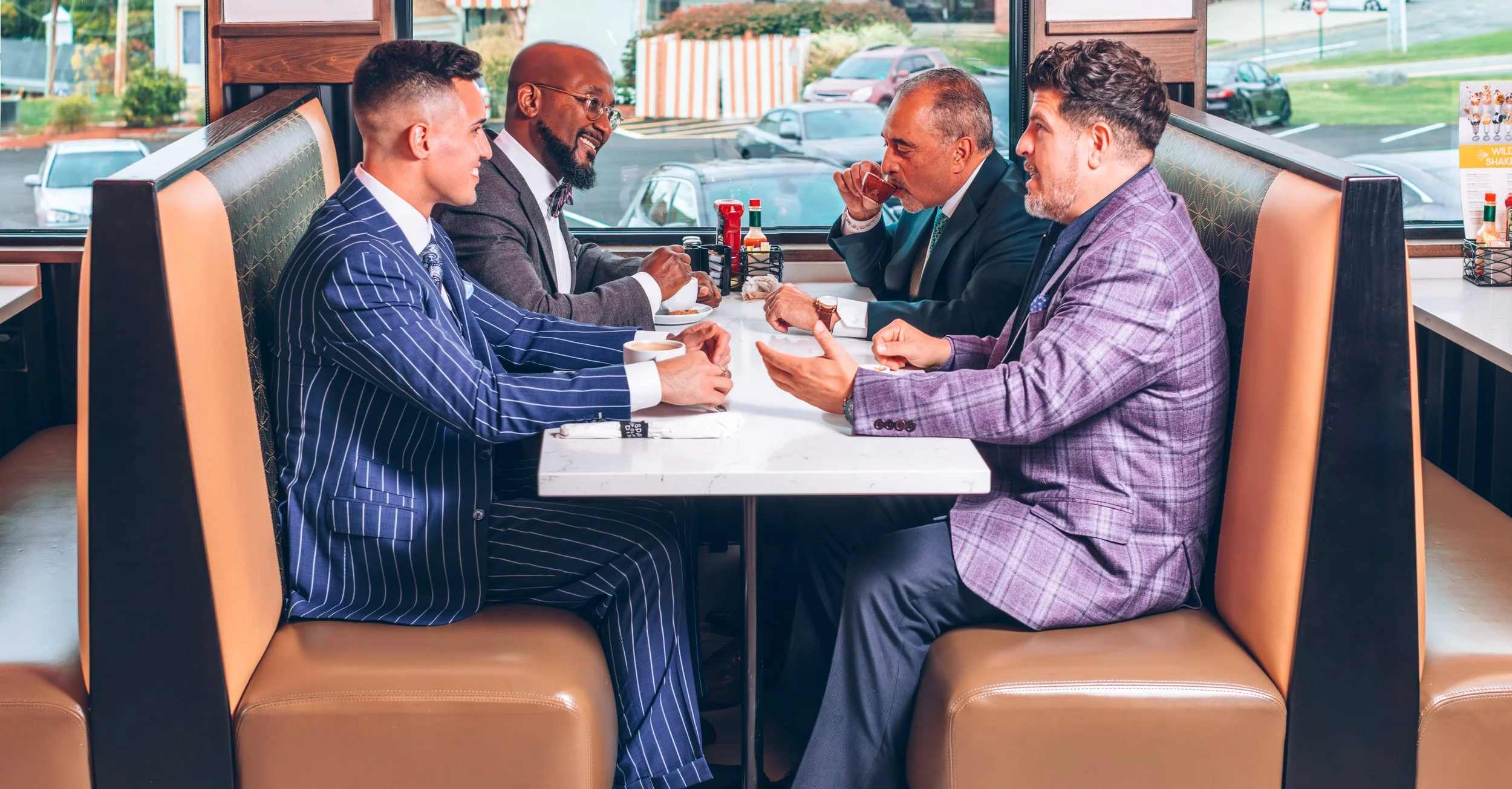 Four men in suits sitting at a restaurant booth, engaged in conversation and smiling, with coffee cups on the table, riding in a casual setting with a window view of parking lots and greenery.