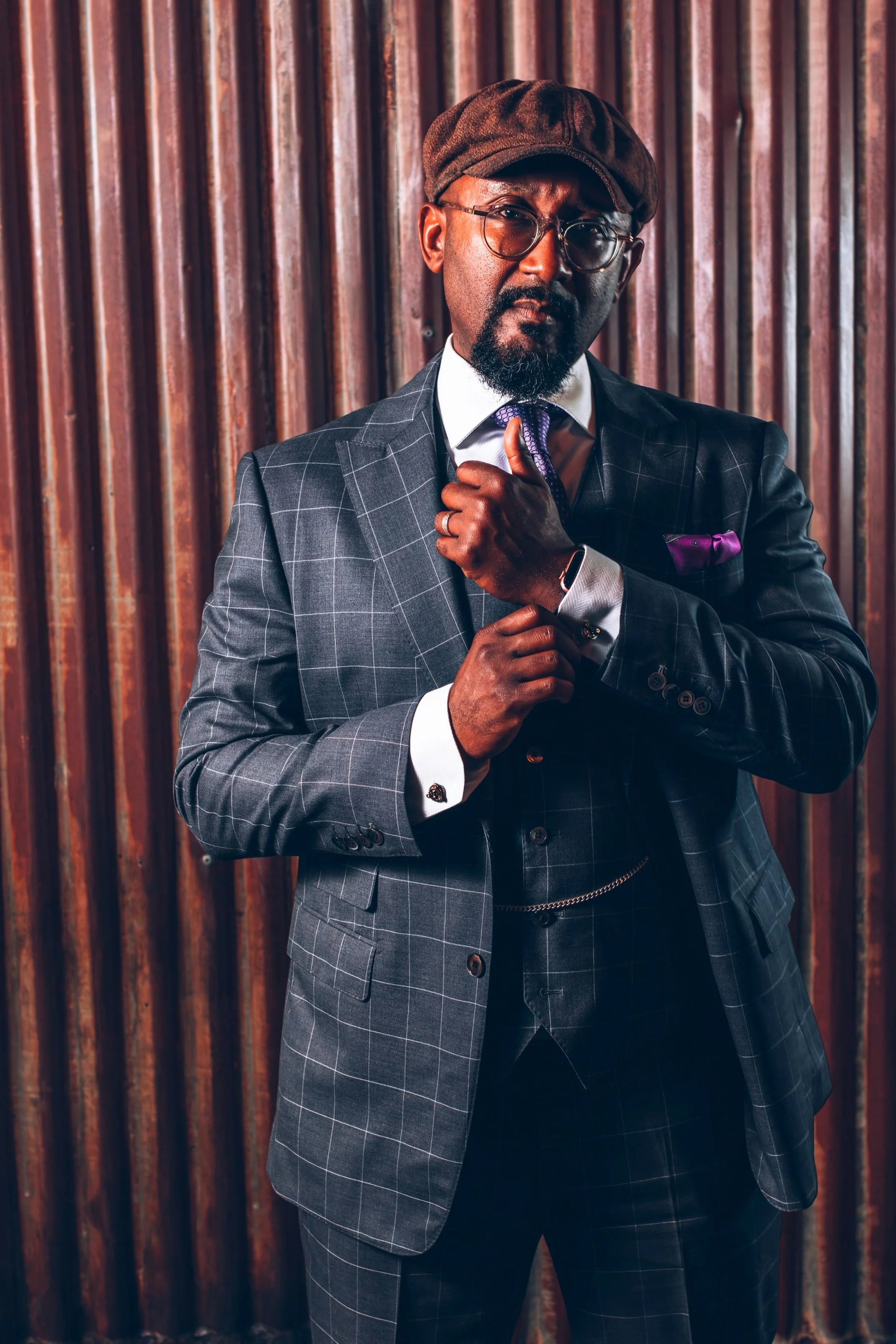 A stylish man dressed in a checked suit with a purple pocket square, wearing glasses and a flat cap, standing against a wooden wall.