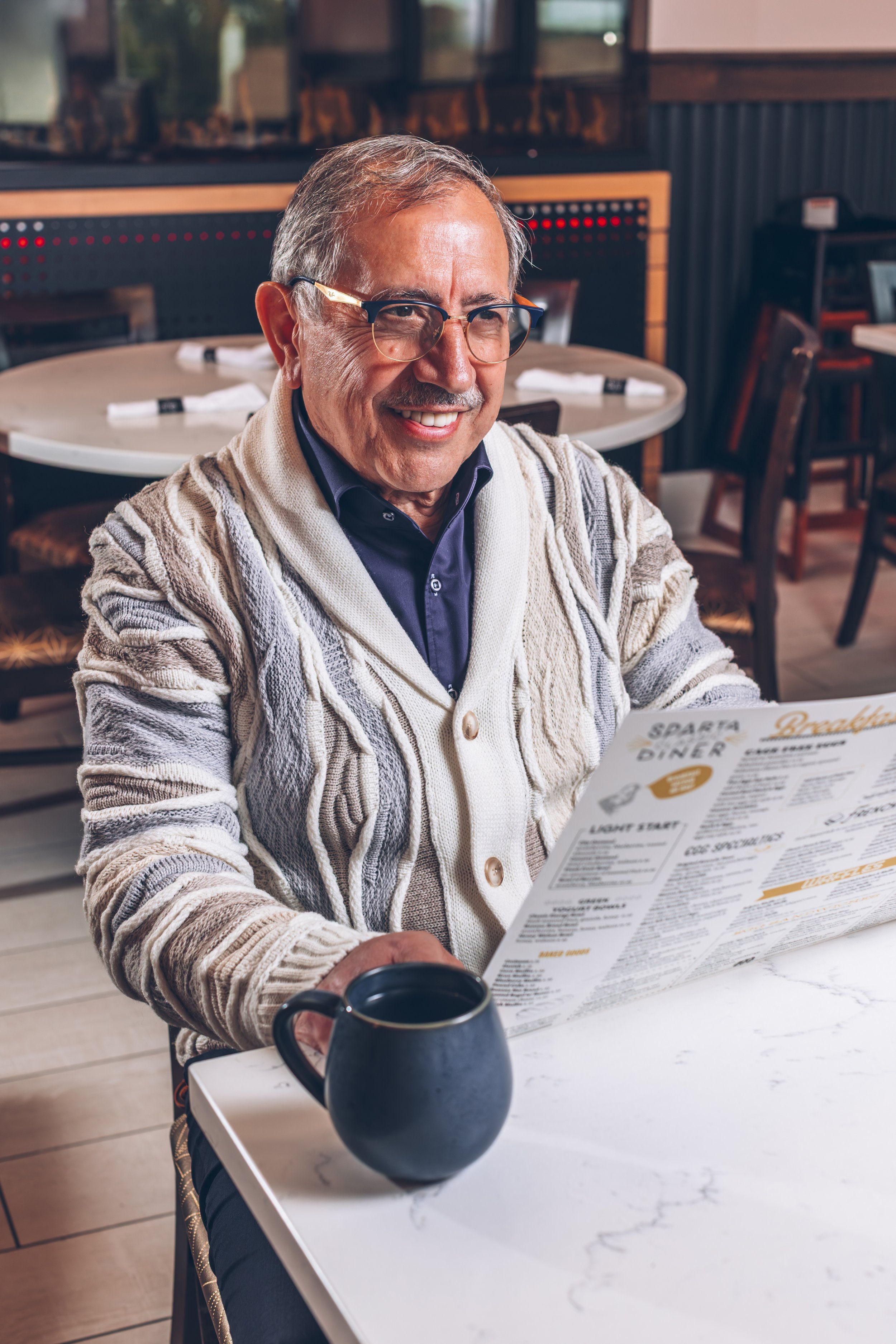 A smiling elderly man with glasses, wearing a patterned cardigan, reading a menu at a restaurant table with a black coffee mug in front of him.