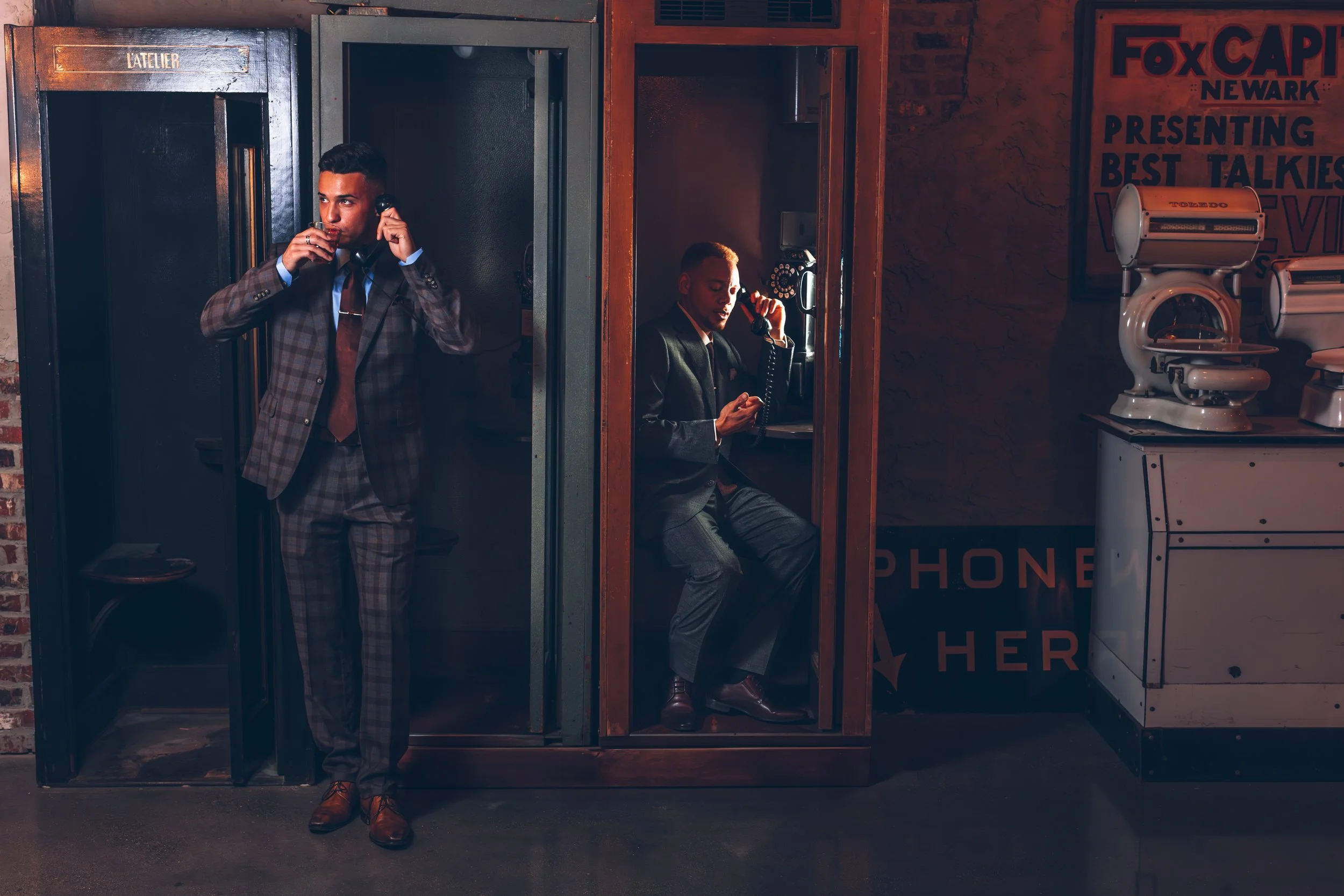 Two men in suits using vintage payphones in a retro-style phone booth, one standing and the other sitting, inside a dimly lit room with old glory and medical machine signage.