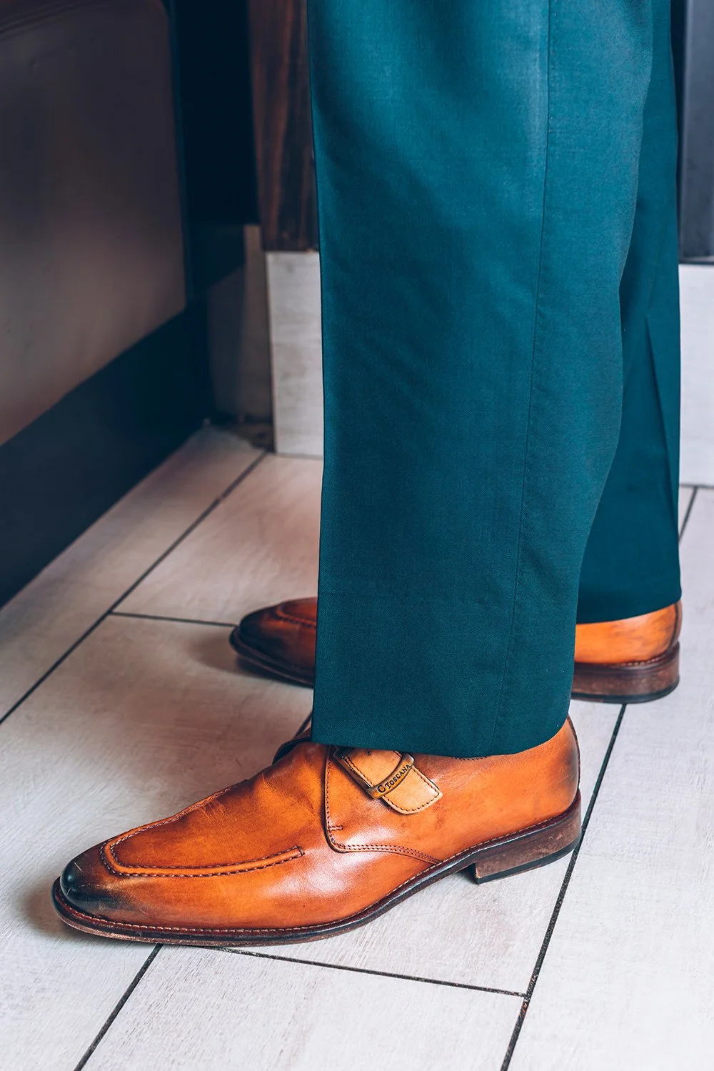 Close-up of brown leather shoes worn with teal pants, standing on tiled floor.