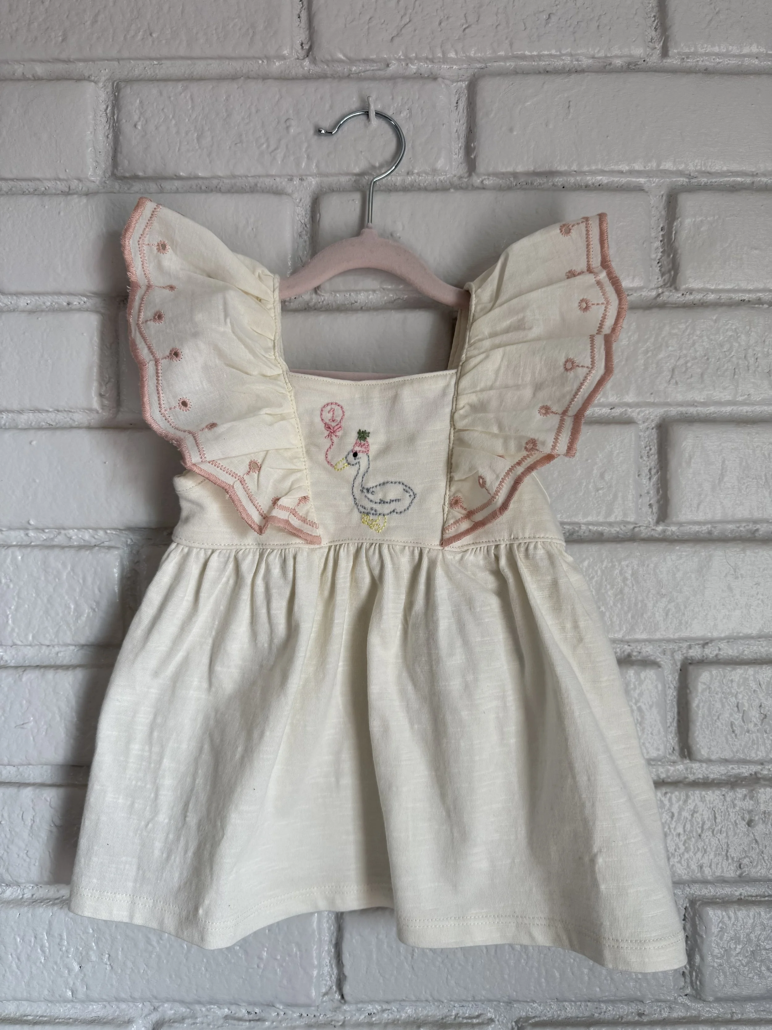 A cream-colored child's dress with embroidered goose and balloon, pink ruffled sleeves with embroidery, hanging on a pastel pink hanger against a white brick wall.