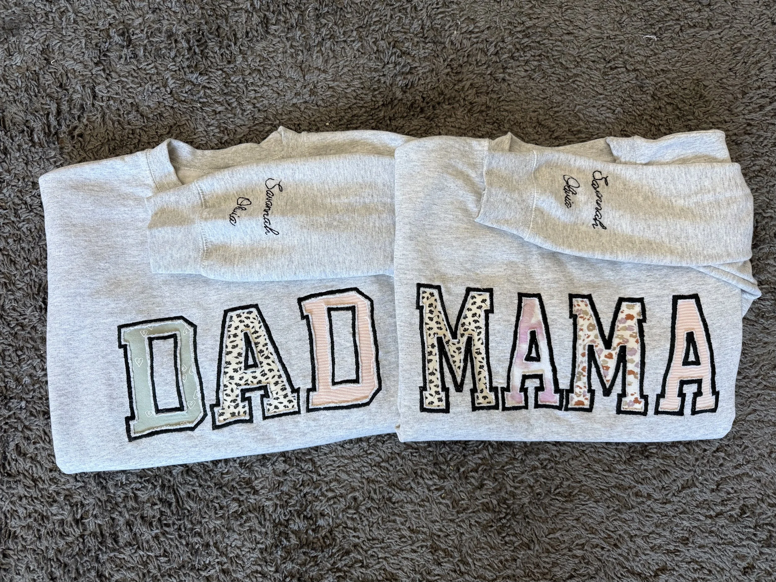Two gray shirts with embroidered text that reads 'Savannah and Olivia' on the sleeve. One shirt has the word 'DAD' with each letter in different pastel colors and patterns, and the other shirt has the words 'MAMA' in similar style.