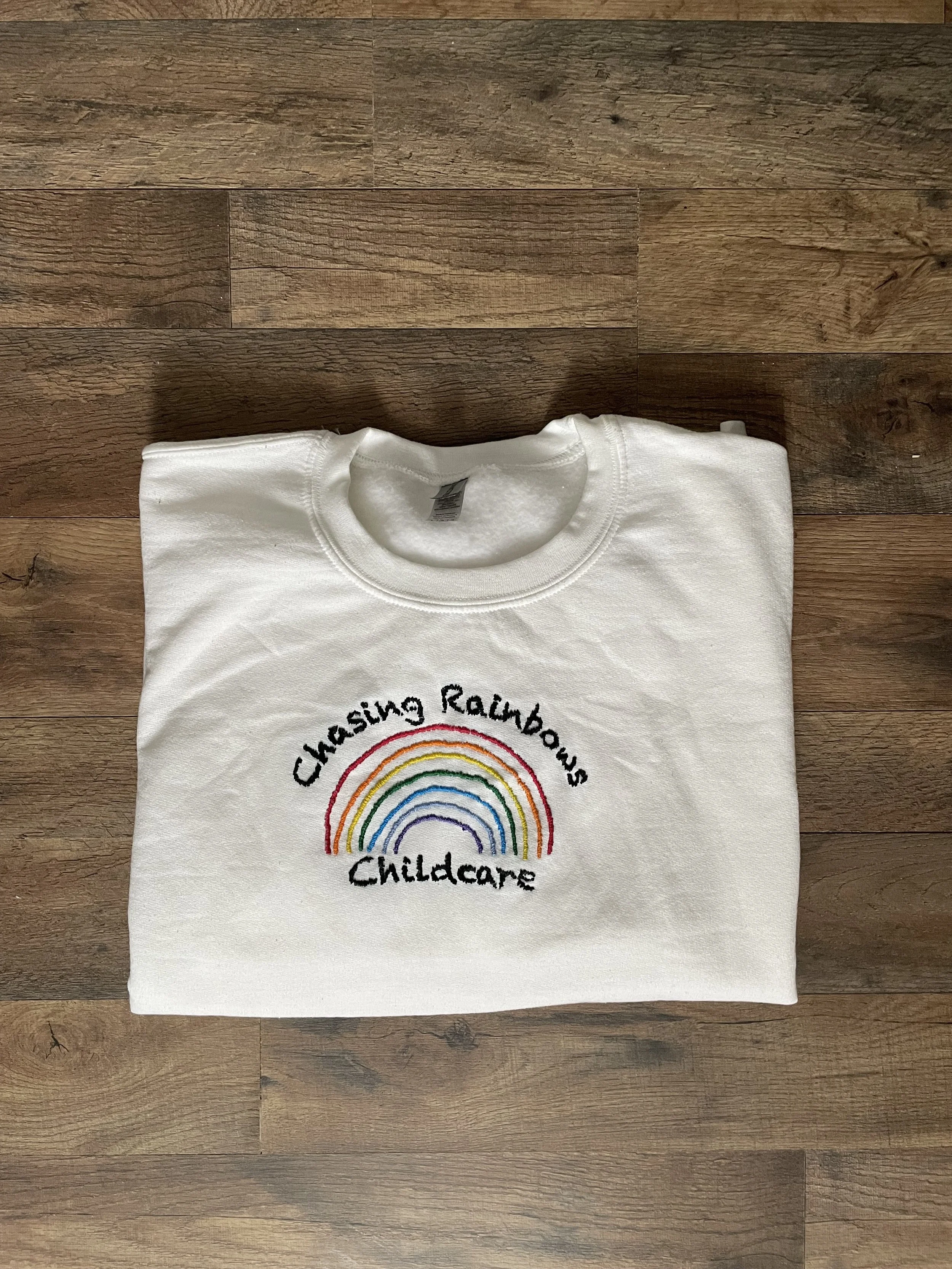 White sweatshirt with a rainbow embroidery and the words 'Chasing Rainbows Childcare'.