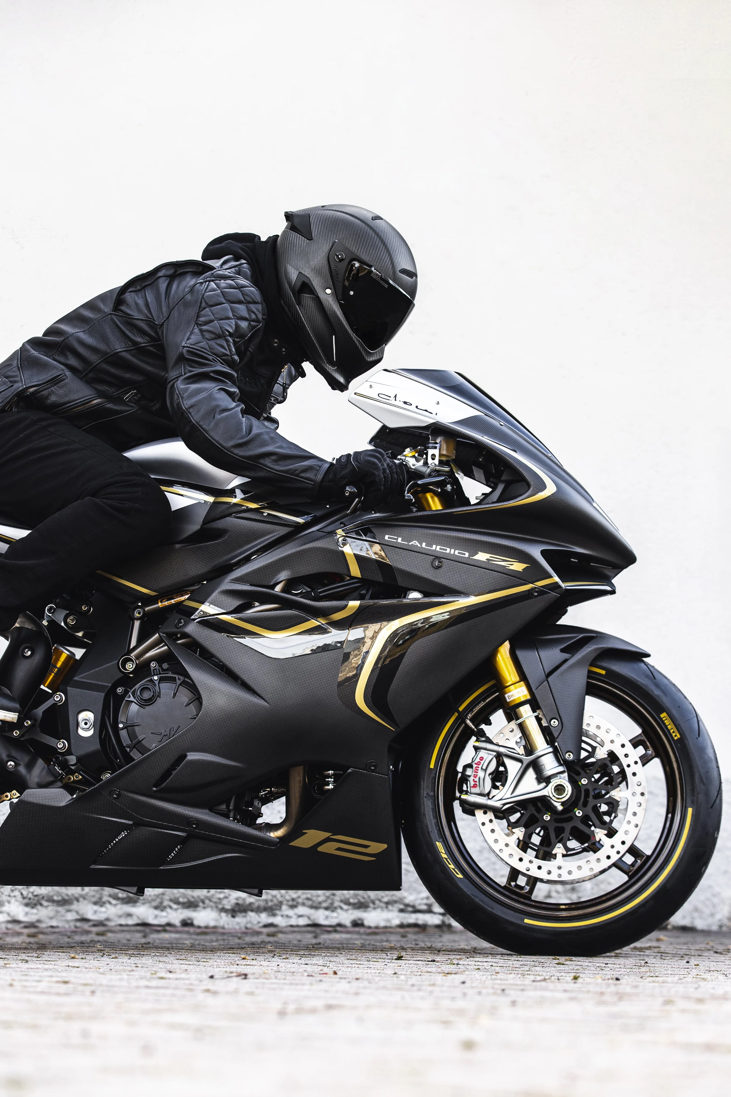 A person wearing a black leather jacket and helmet sitting on a black and gold sport motorcycle against a plain white wall.