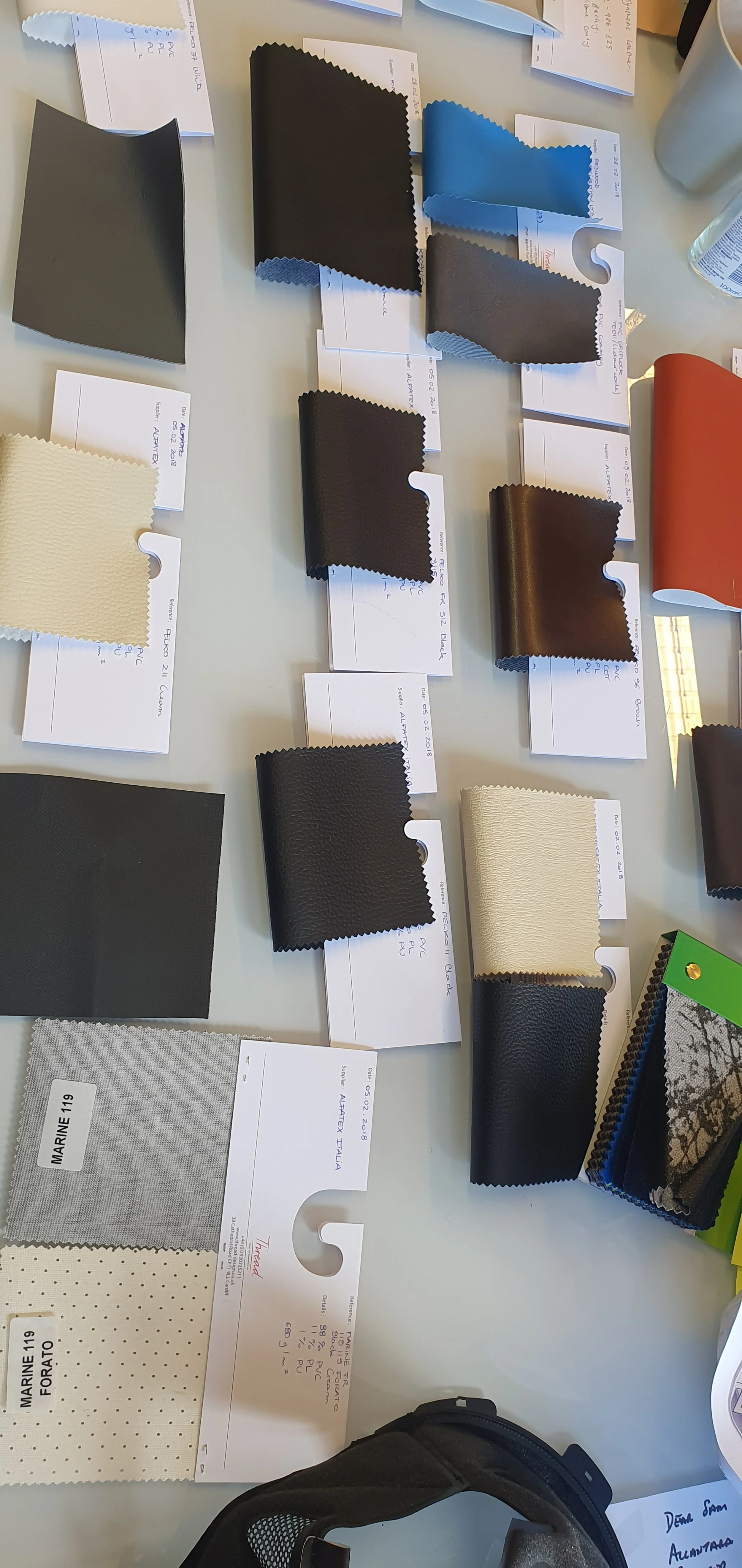 Several fabric swatches in different colors and textures laid out on a table with handwritten notes beneath each sample, alongside some paper and a bag.