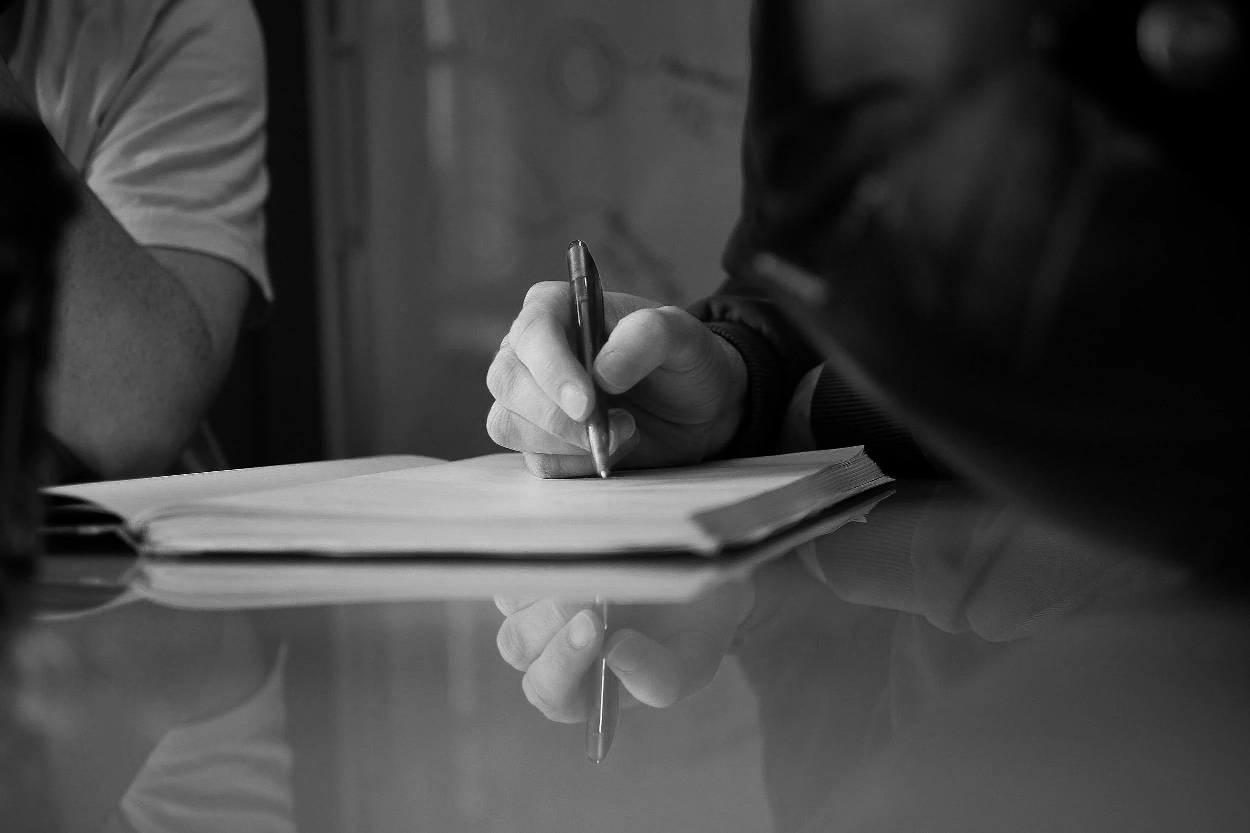 Close-up of a person writing on a notepad with a pen, seated at a reflective surface, in black and white.