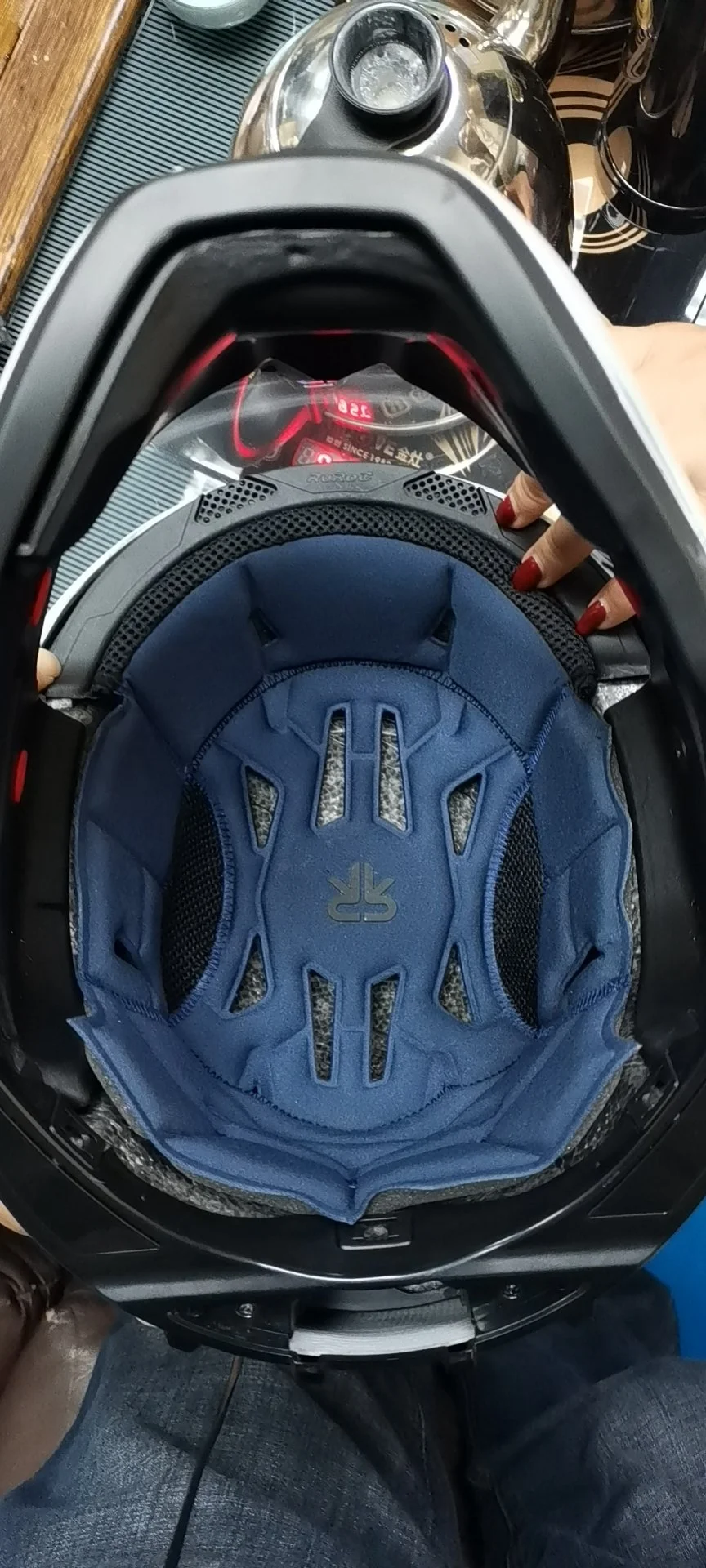 View from above of a motorcycle helmet with the blue interior lining visible, held by a person with red nails, with a kitchen counter and kettle in the background.