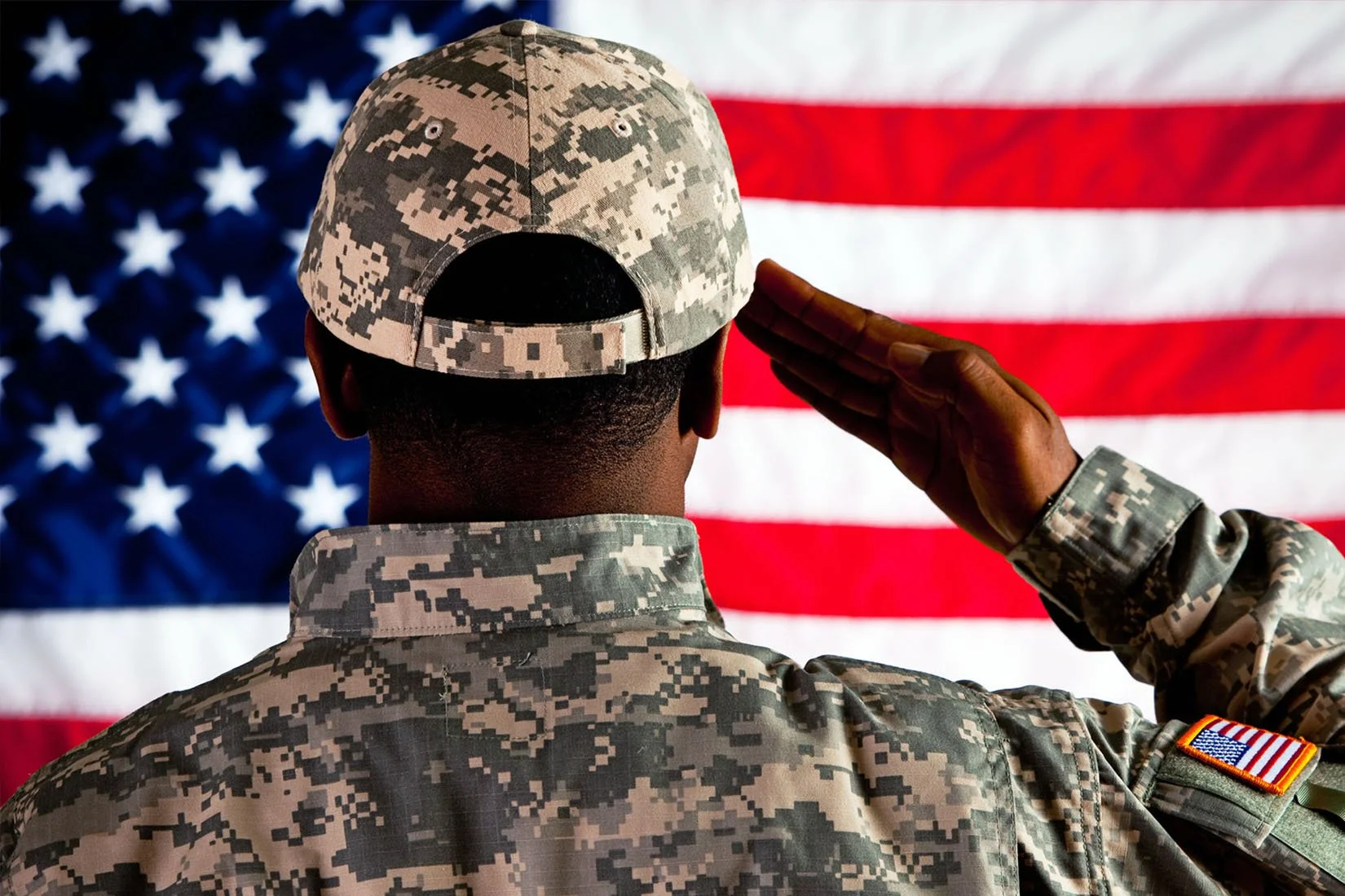 A person in military camouflage uniform salutes in front of an American flag background.