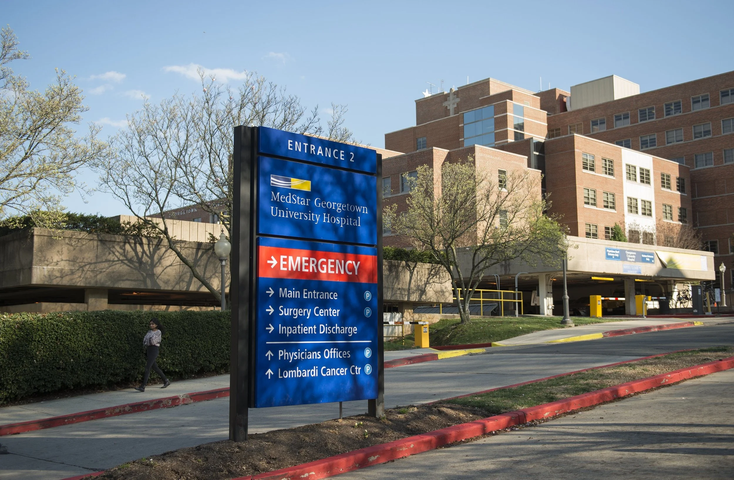 Signage outside MedStar Georgetown University Hospital indicating Entrance 2 and various departments including emergency, main entrance, surgery center, inpatient discharge, physicians offices, and Lombardi Cancer Center. A brick hospital building and leafless trees are visible in the background under a clear blue sky.