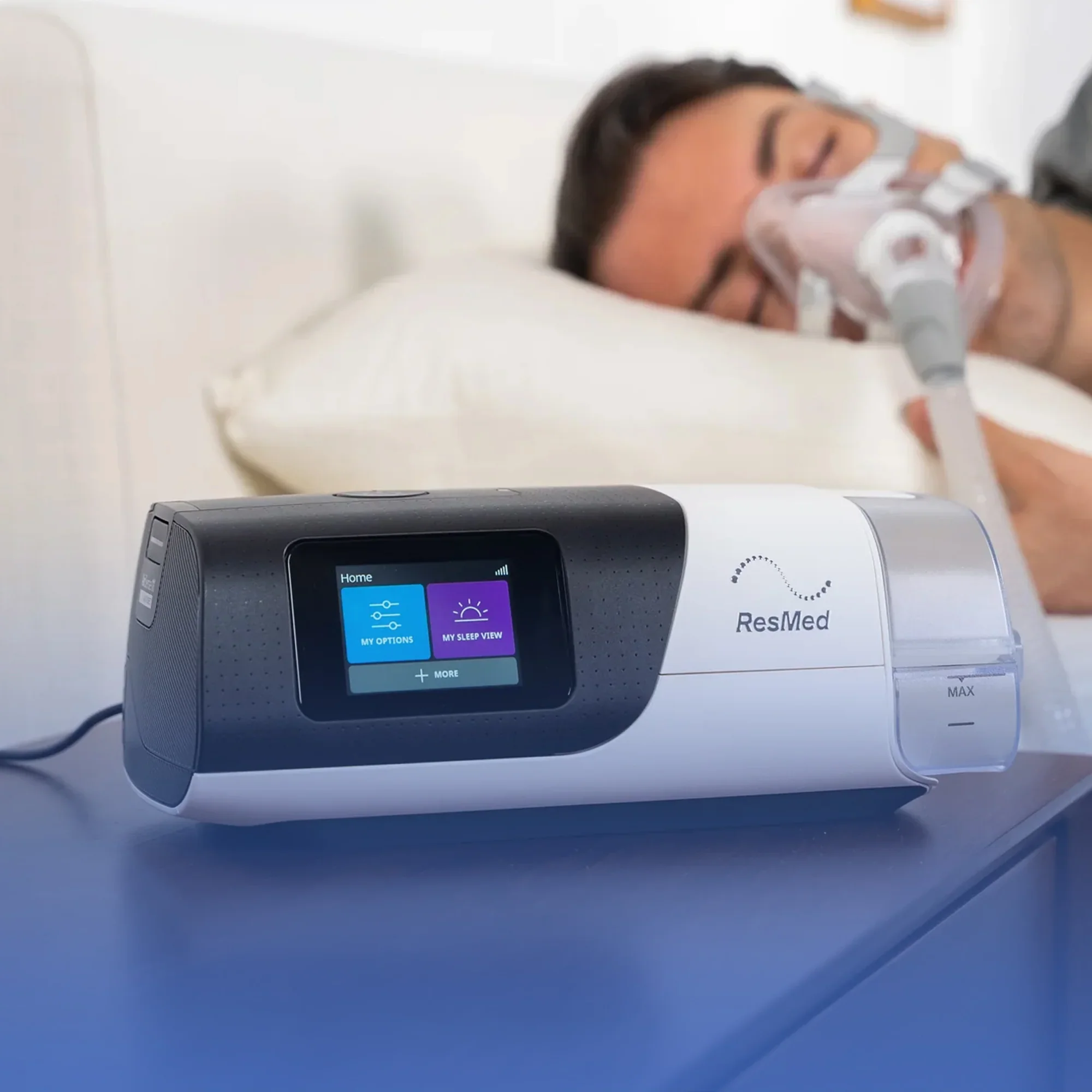 Close-up of a white ResMed CPAP machine with a digital display screen showing options for sleep therapy, placed on a blue surface with a white pillow in the background.