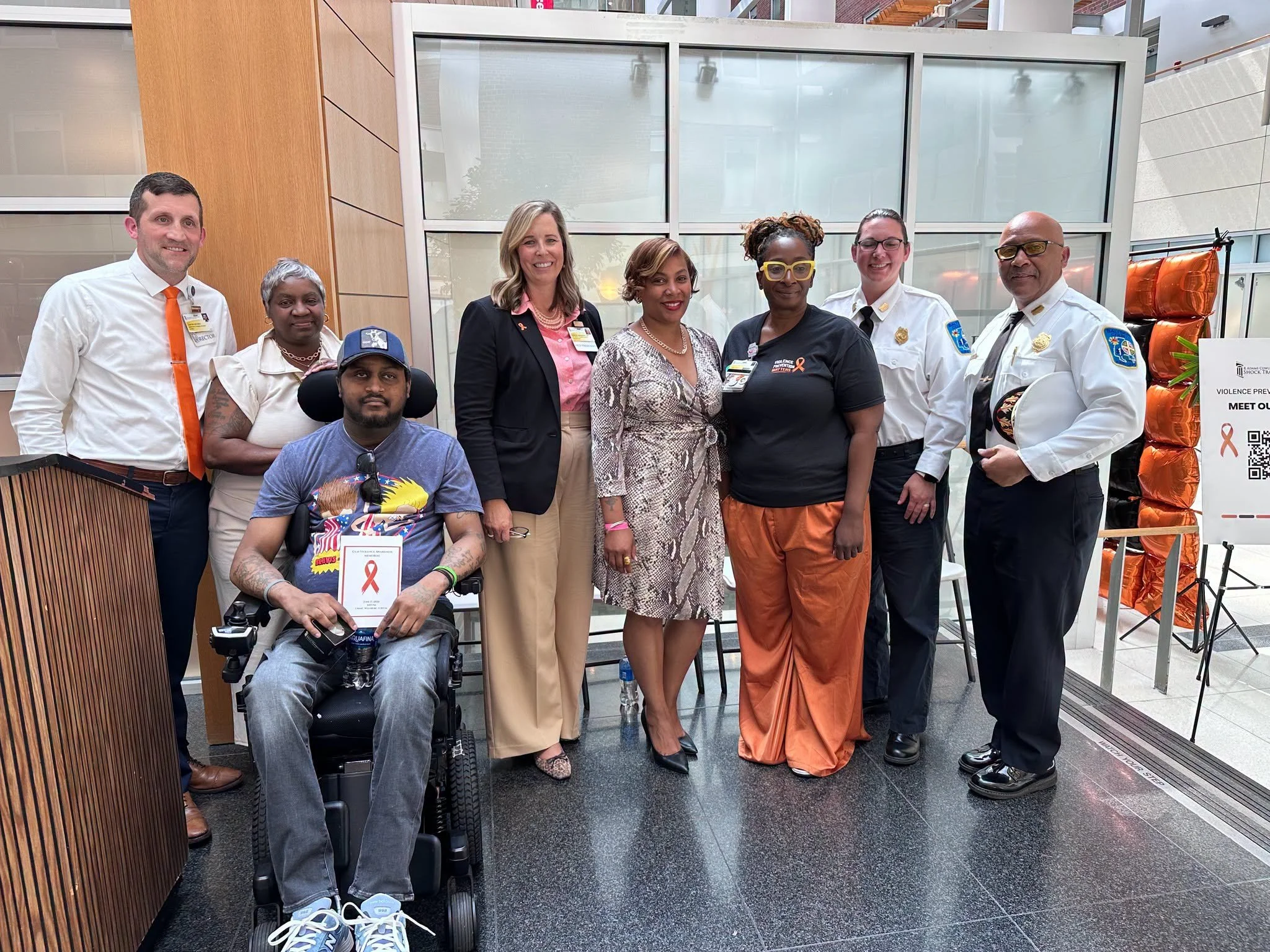 A diverse group of nine people standing and sitting together indoors, some wearing official uniforms, in front of a large glass window, with a sign to the right about violence prevention awareness.