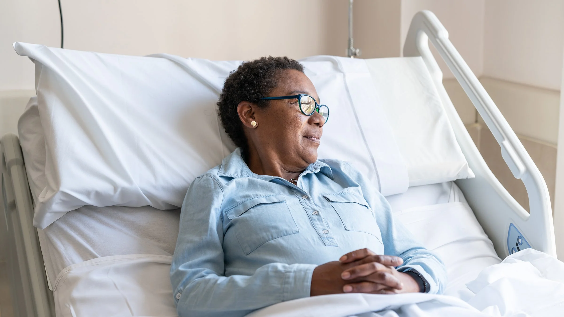 An older woman with short curly hair, glasses, and earrings resting in a hospital bed with white sheets and pillow, wearing a light blue button-up shirt, looking to the side with a calm expression.