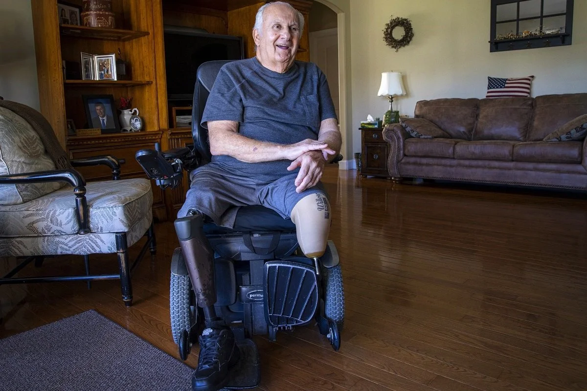 An elderly man with a prosthetic leg sitting in a motorized wheelchair in a living room, smiling and looking to his right.