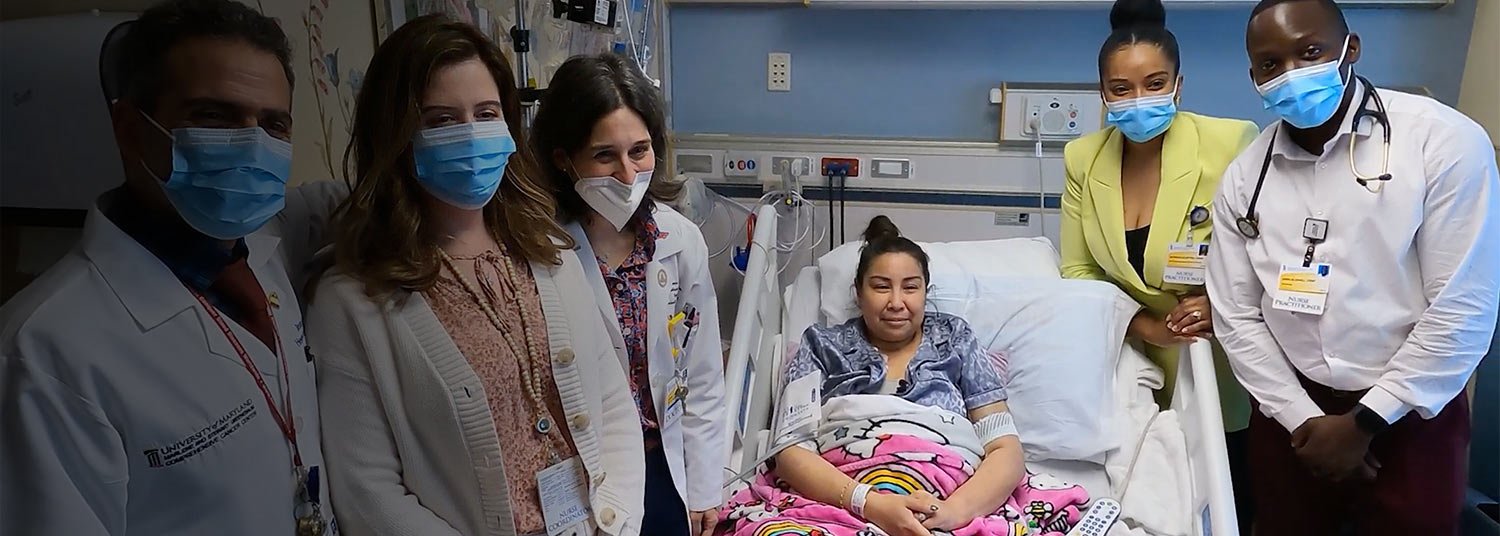 Six healthcare professionals, all wearing face masks, stand around a young girl patient in a hospital bed. The girl, with a cheerful blanket over her lap, is inside a hospital room, smiling at the camera.