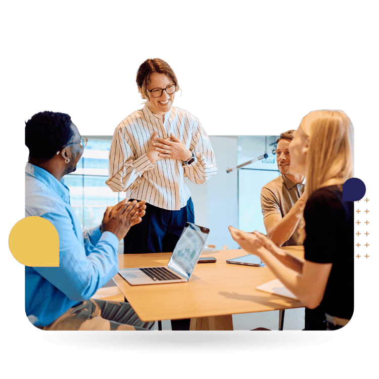 Four colleagues engage in a positive workplace discussion around a conference table, with one person standing and smiling while others applaud