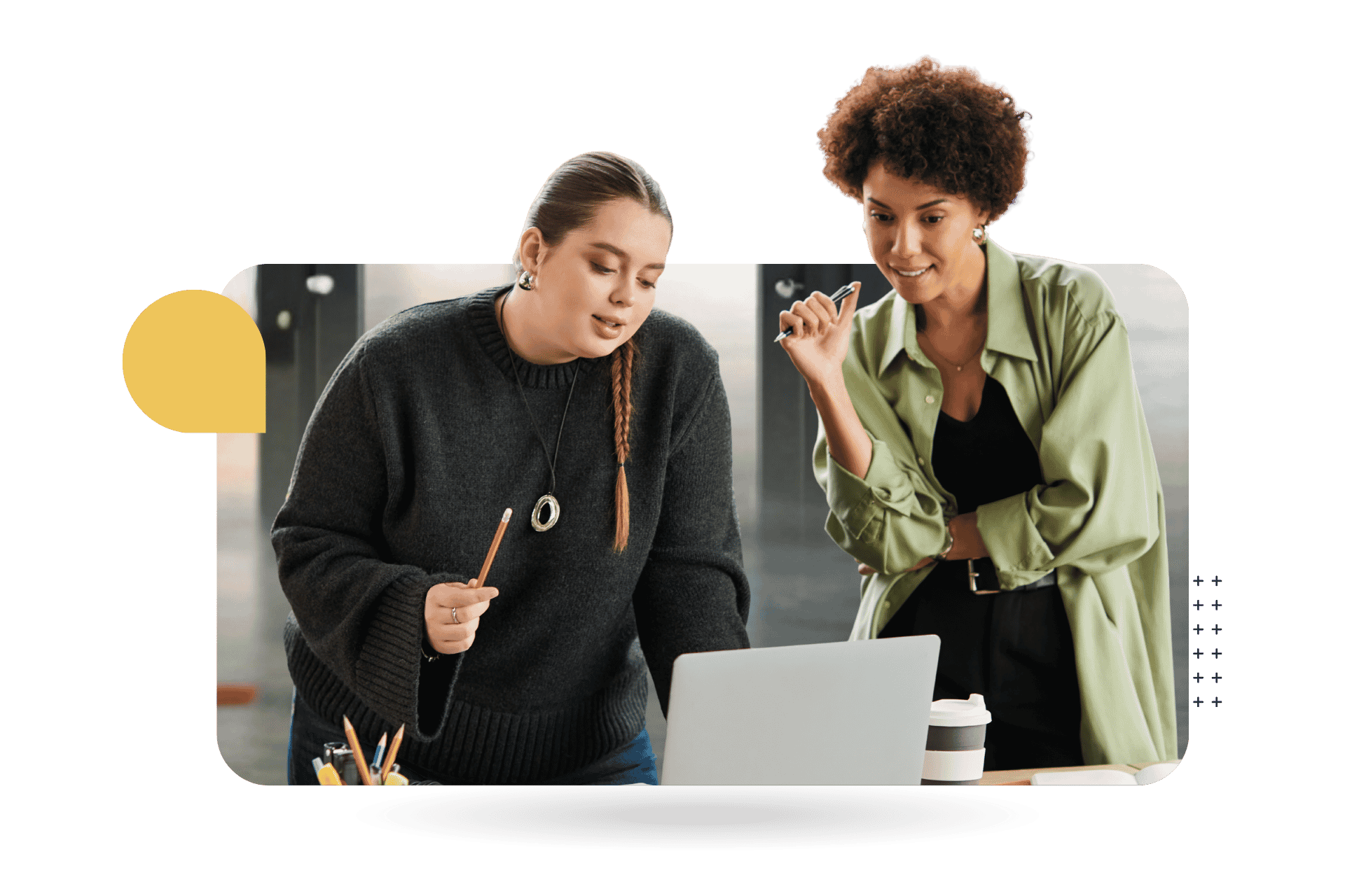 Two women collaborate over a laptop in an office setting, with one pointing at the screen while the other holds a pen.