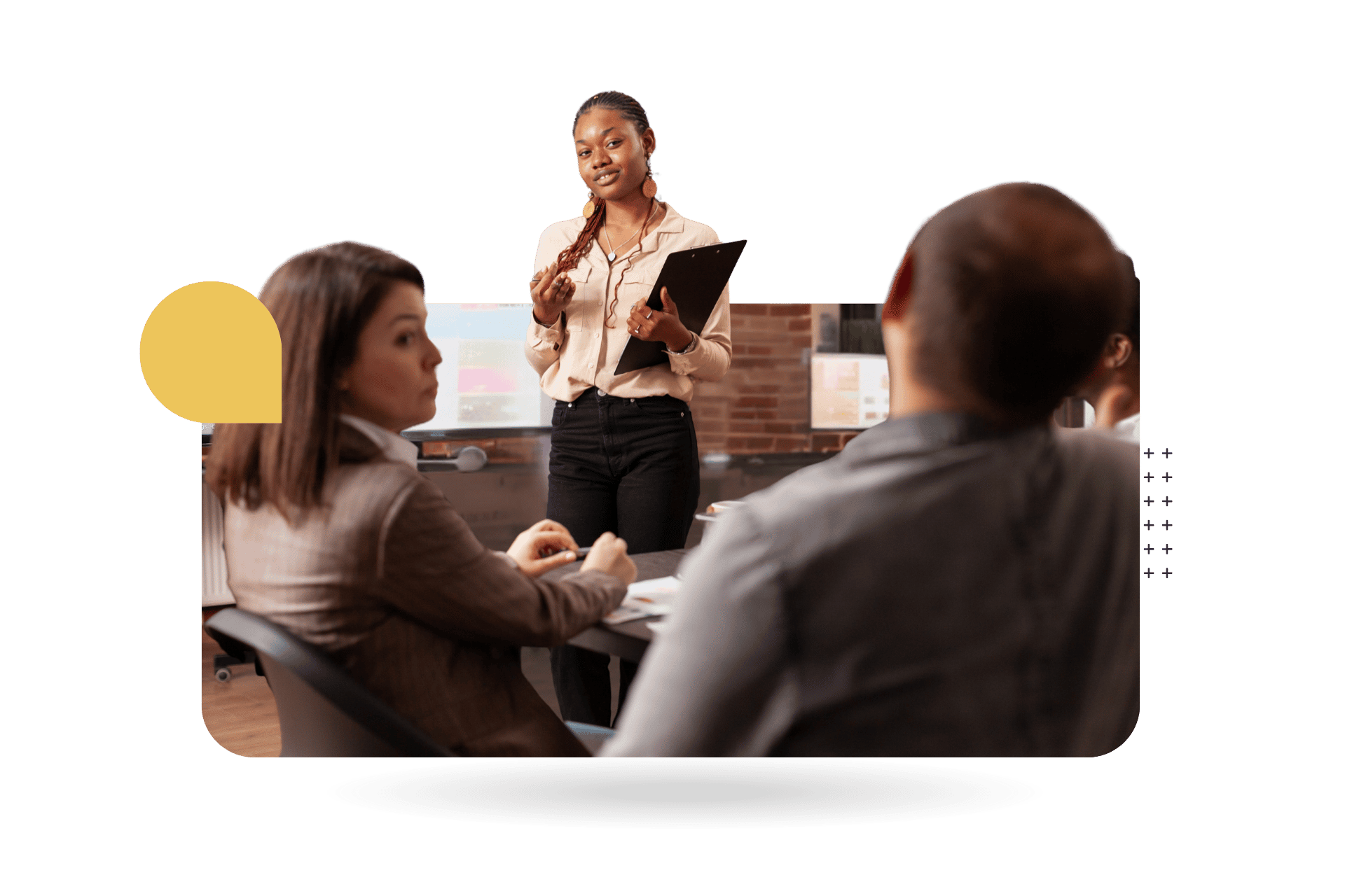 A woman stands at the front of a meeting room holding a clipboard and speaking to a small group seated around a conference table.