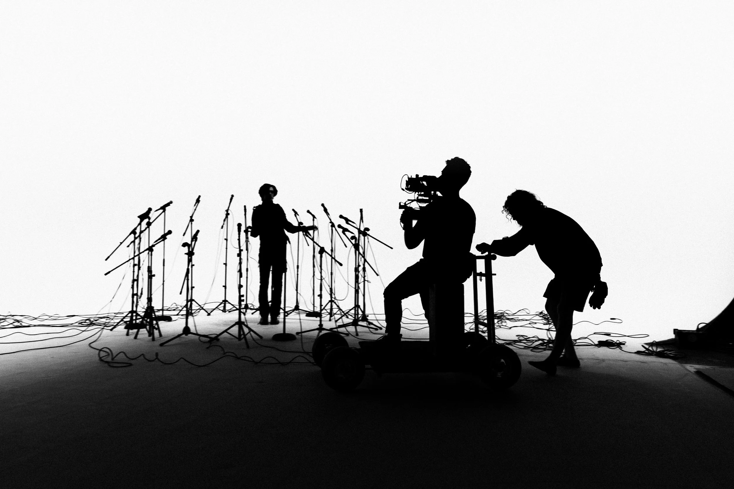 Silhouettes of crew members setting up microphones and equipment on stage with a person filming against a plain background.