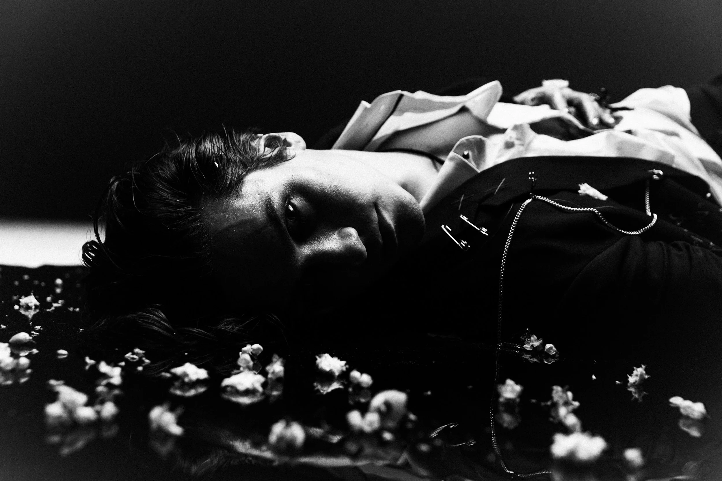 Black and white photo of a person lying on the ground, surrounded by scattered popcorn or similar snack food. The person has medium-length hair and is wearing a jacket with visible zippers and chains.