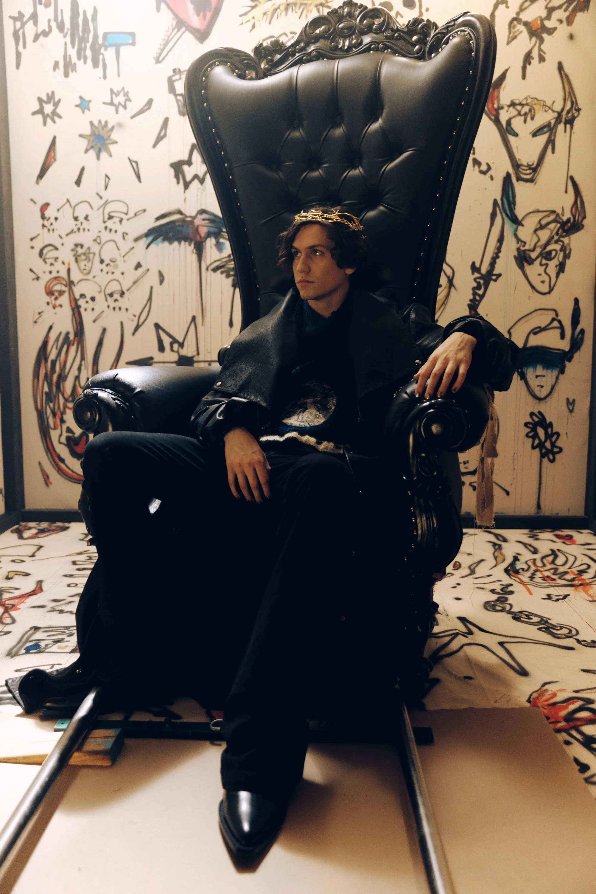 A young man with dark hair sitting on an ornate black leather throne with a high backrest, in front of a wall decorated with colorful graffiti-style artwork including faces, skulls, stars, and abstract shapes, wearing a black leather jacket and black