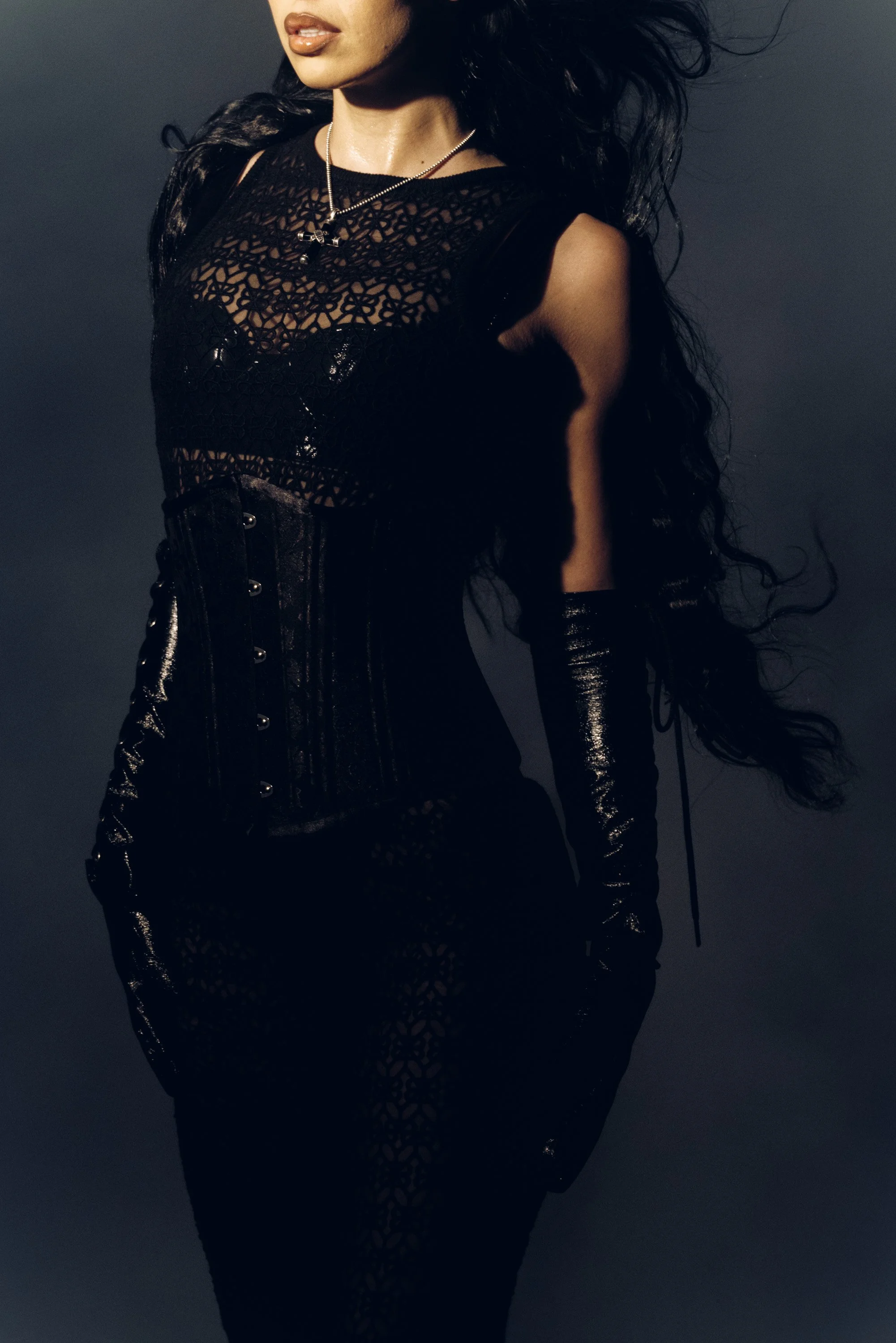 A woman with dark, wavy hair wearing a black lace top with intricate floral patterns, paired with shiny black high-waisted pants, also with a lace-up detail. She is accessorized with a silver necklace and long, black gloves, set against a dark backgr