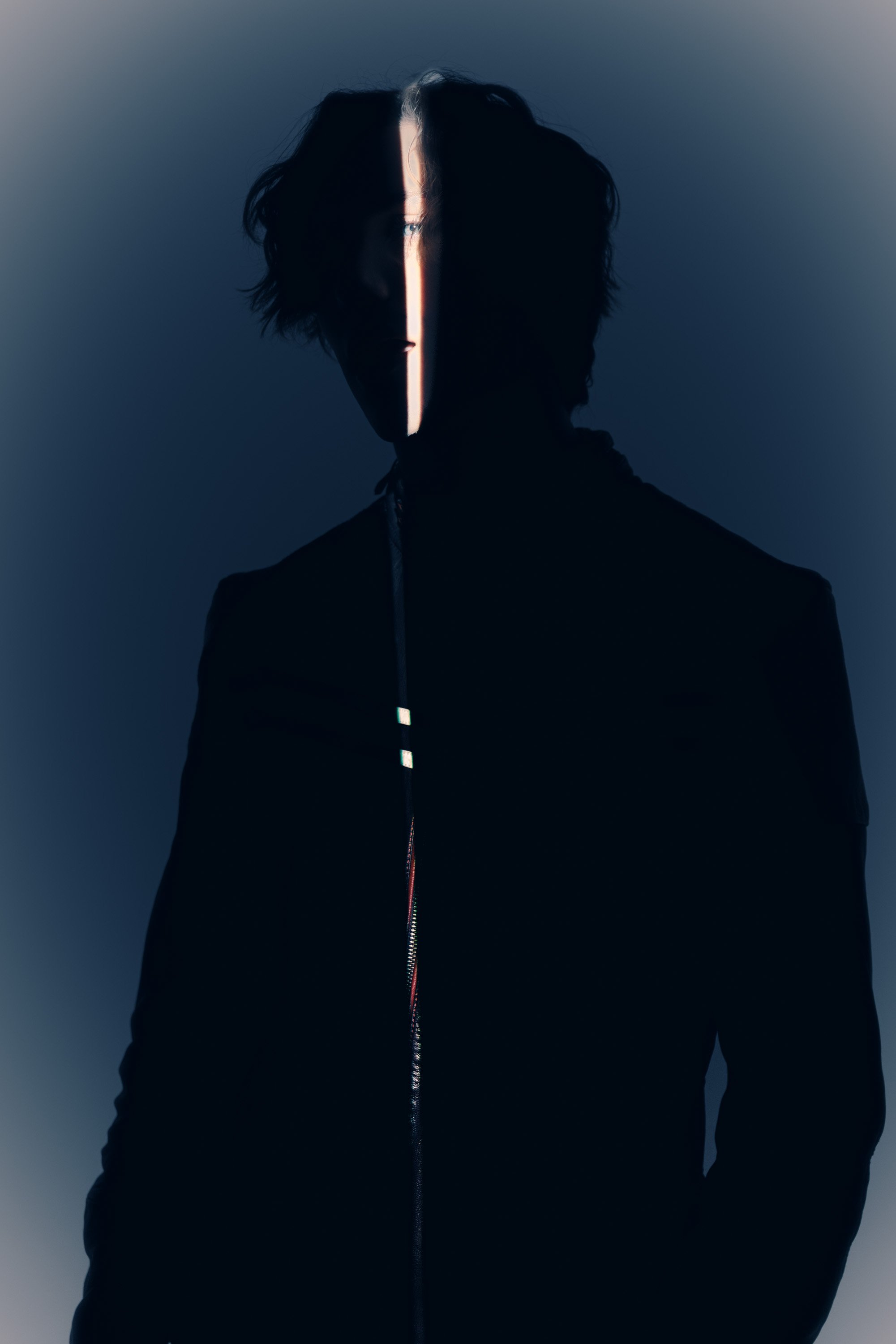 A person with curly hair is wearing a black jacket with a zipper, and a vertical strip of light runs from the top to the bottom of the image, illuminating parts of their face and chest.