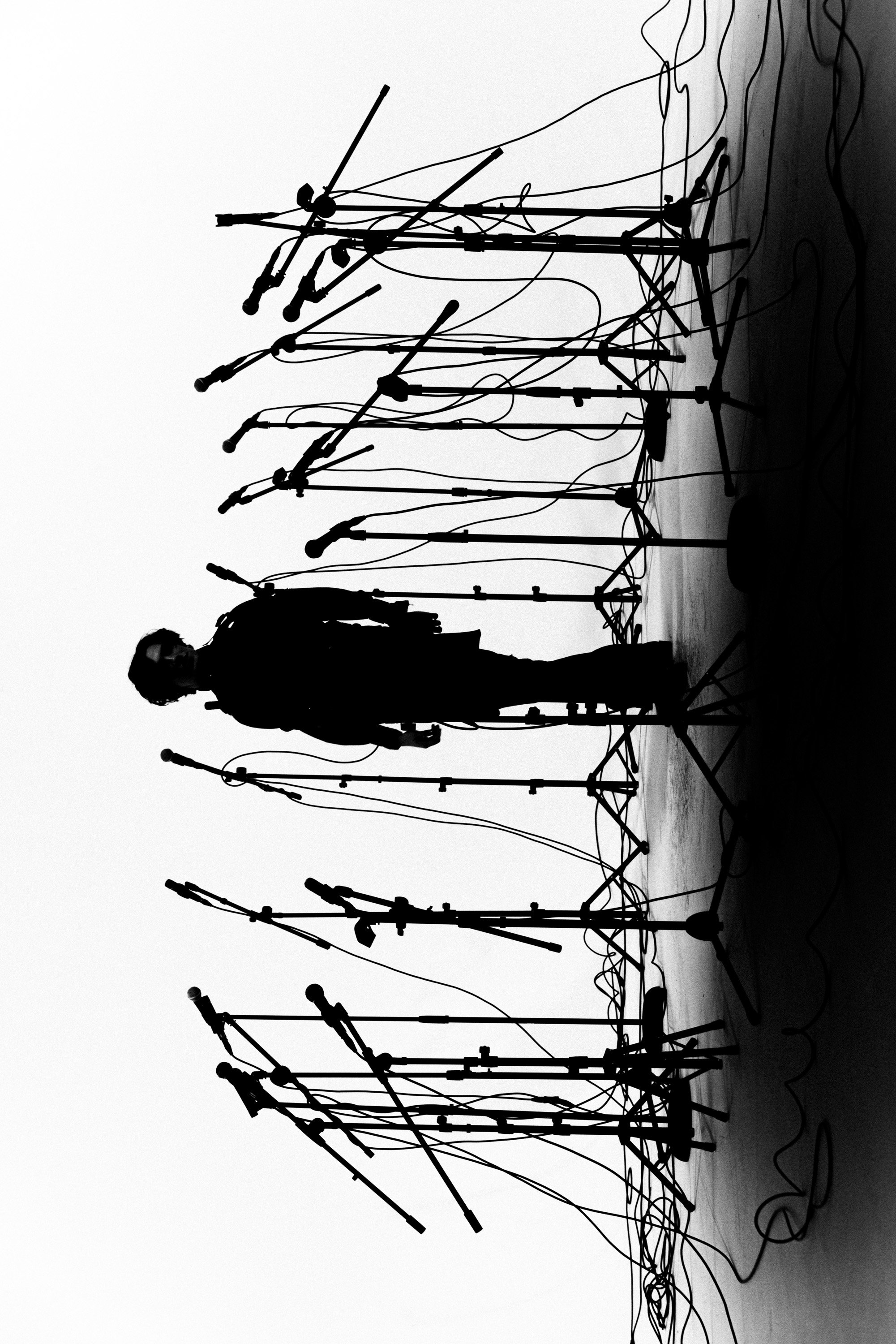 Black and white photo of a person with curly hair and glasses standing behind a large number of microphones on stands, arranged in multiple rows and angles, likely in a recording studio or a press conference setting.