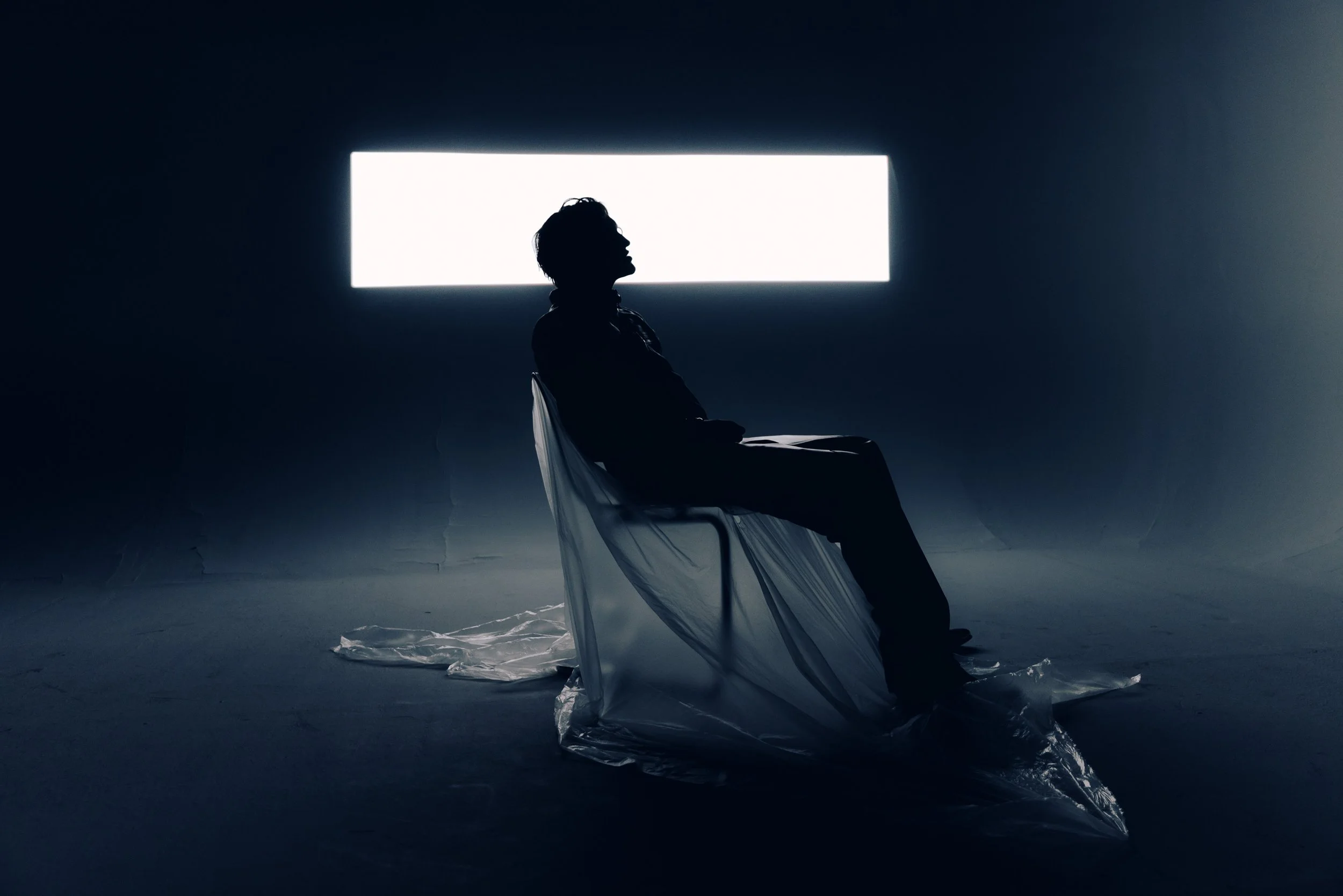 Silhouette of a person seated on a chair in front of a bright rectangular light source in a dark room, with a crumpled plastic sheet on the floor.