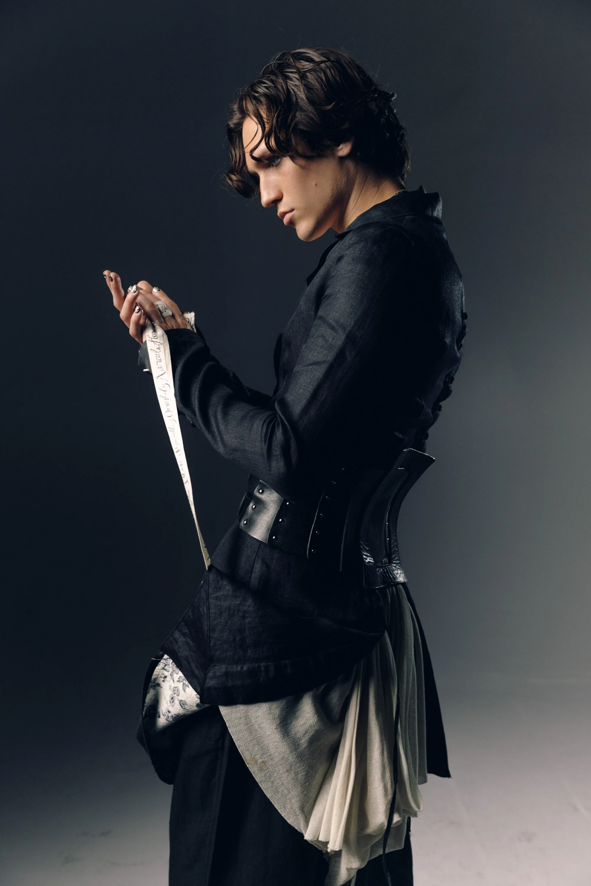 A person with medium-length wavy hair and fair skin, dressed in dark clothing, is looking down at their hands holding a measuring tape against a dark background.