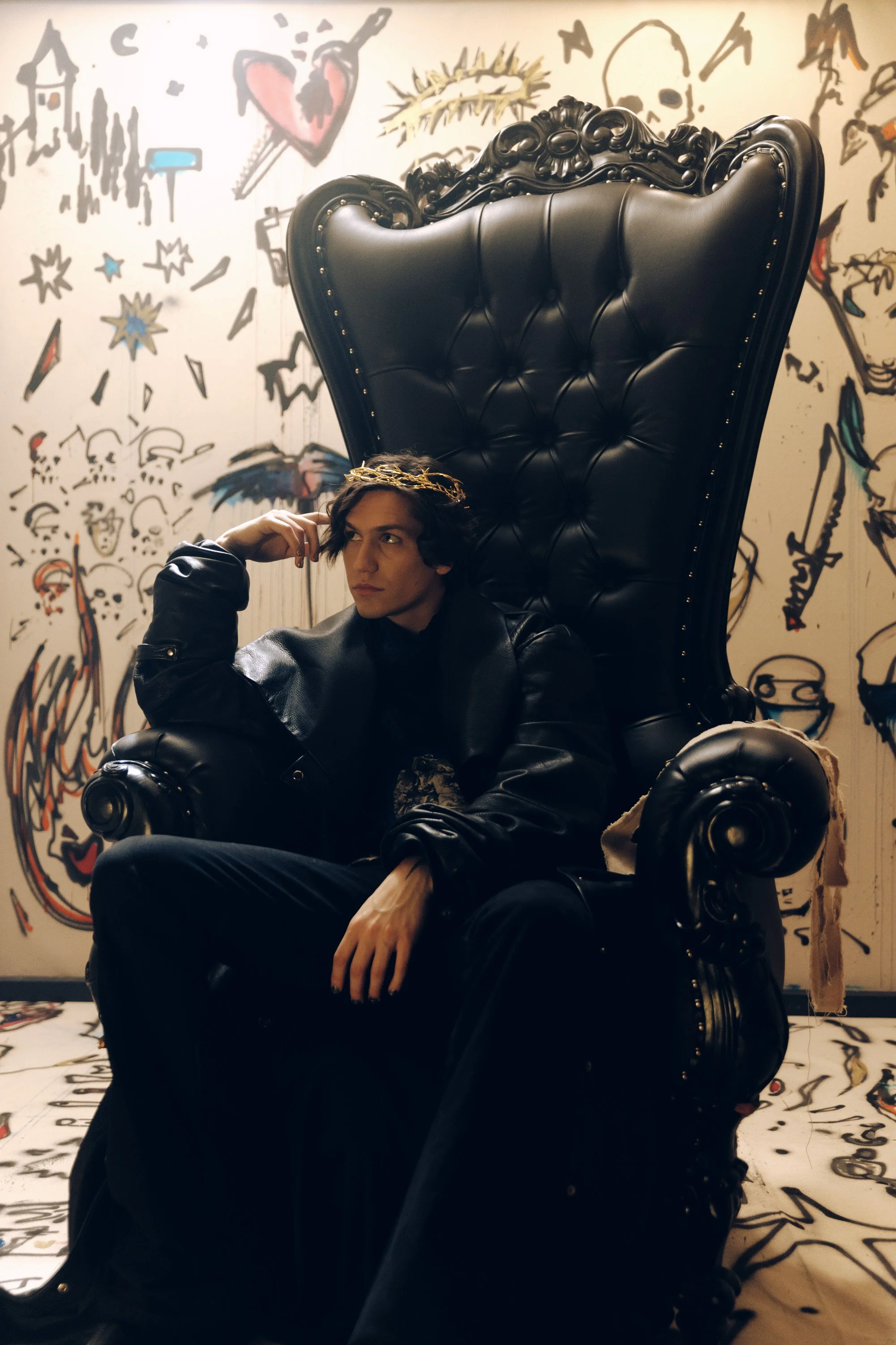 Young man with dark hair wearing a black leather jacket and dark pants, sitting on a large black throne-like chair with ornate carved wood details, in front of a graffiti wall with various drawings including a heart with a dagger, skulls, and other a