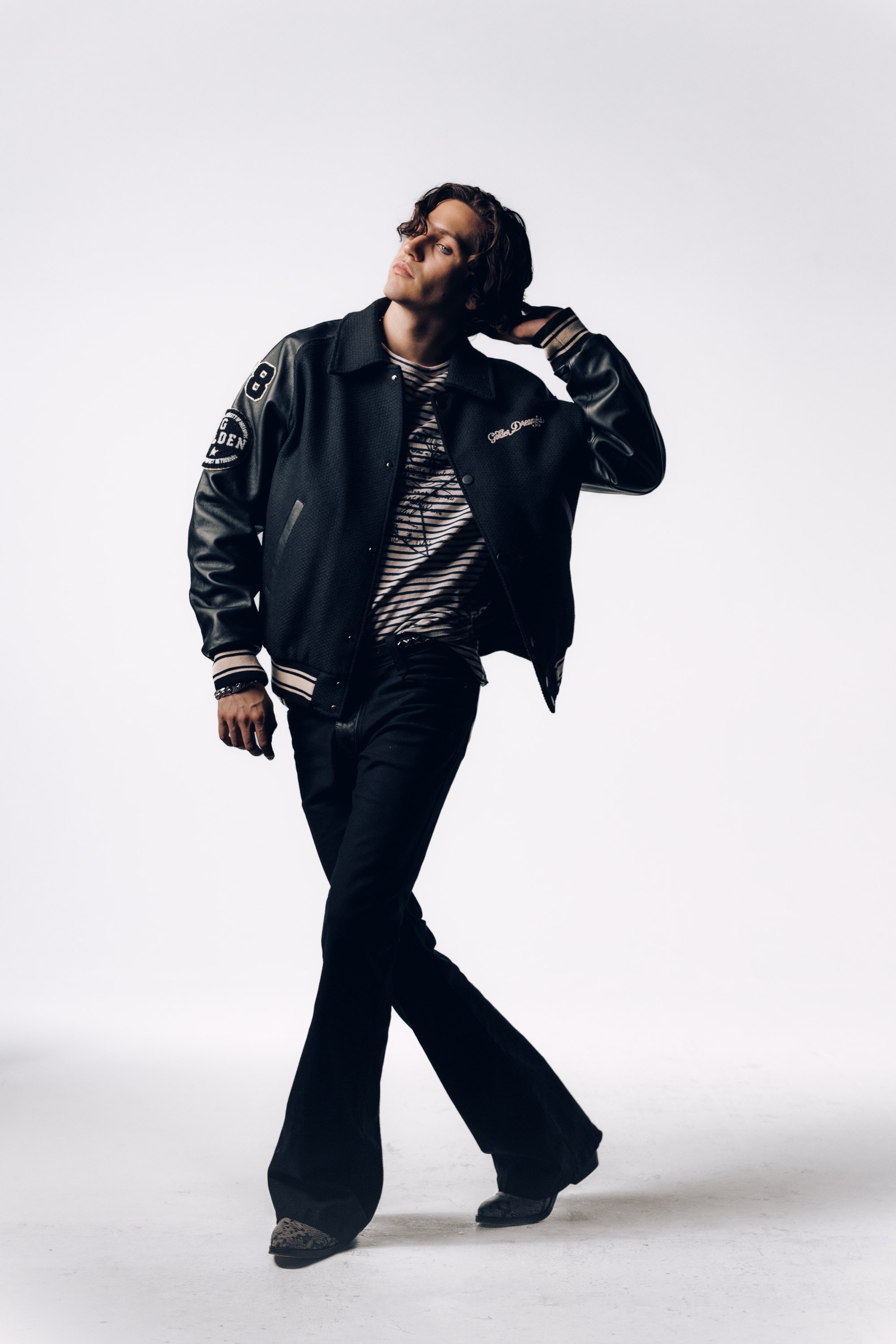 A person stands with a pose against a plain white background. They wear a black varsity jacket, striped shirt, and wide-leg black pants.