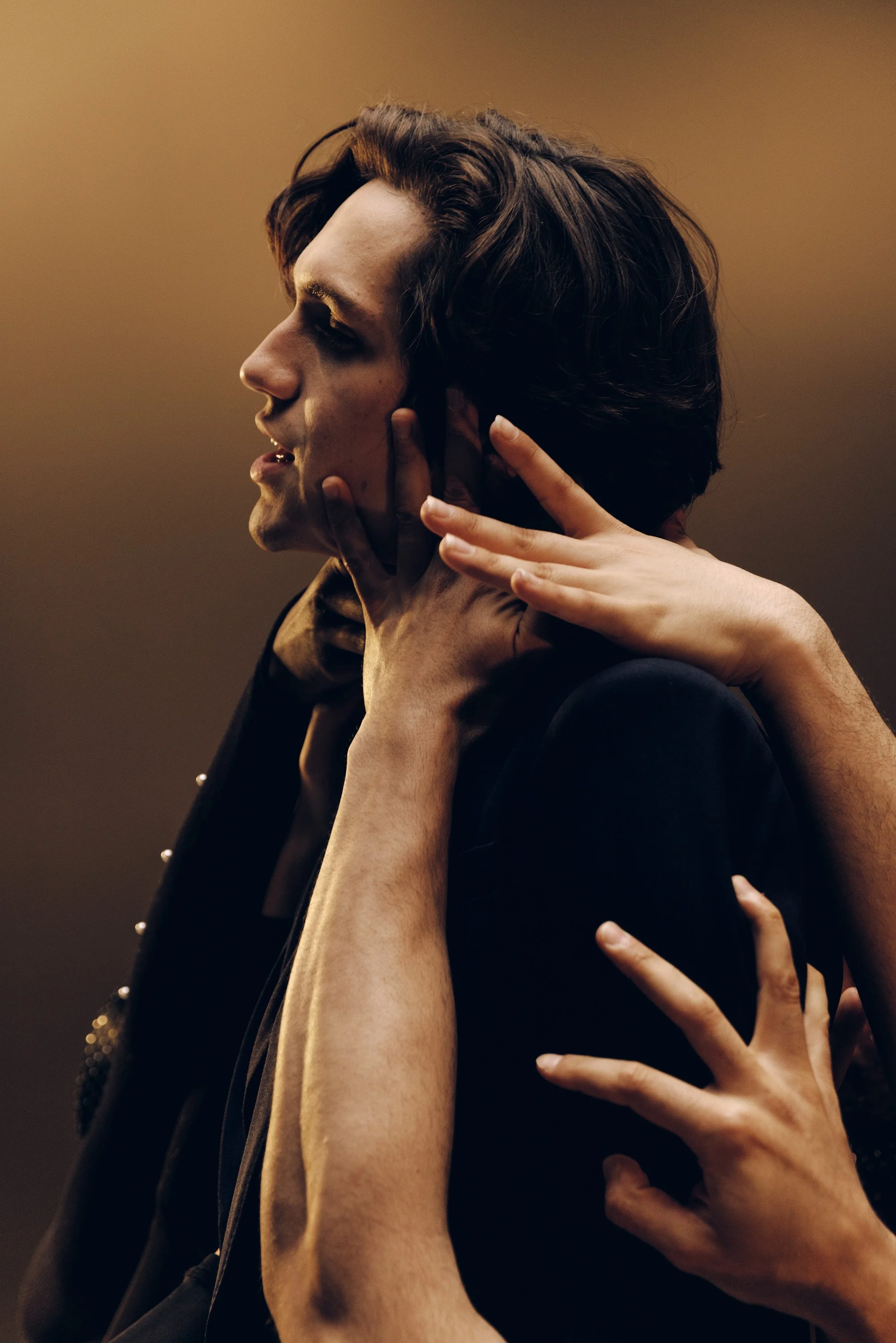 Close-up of a person with dark hair and makeup, being touched gently on the face and neck by hands, with a warm brown background.