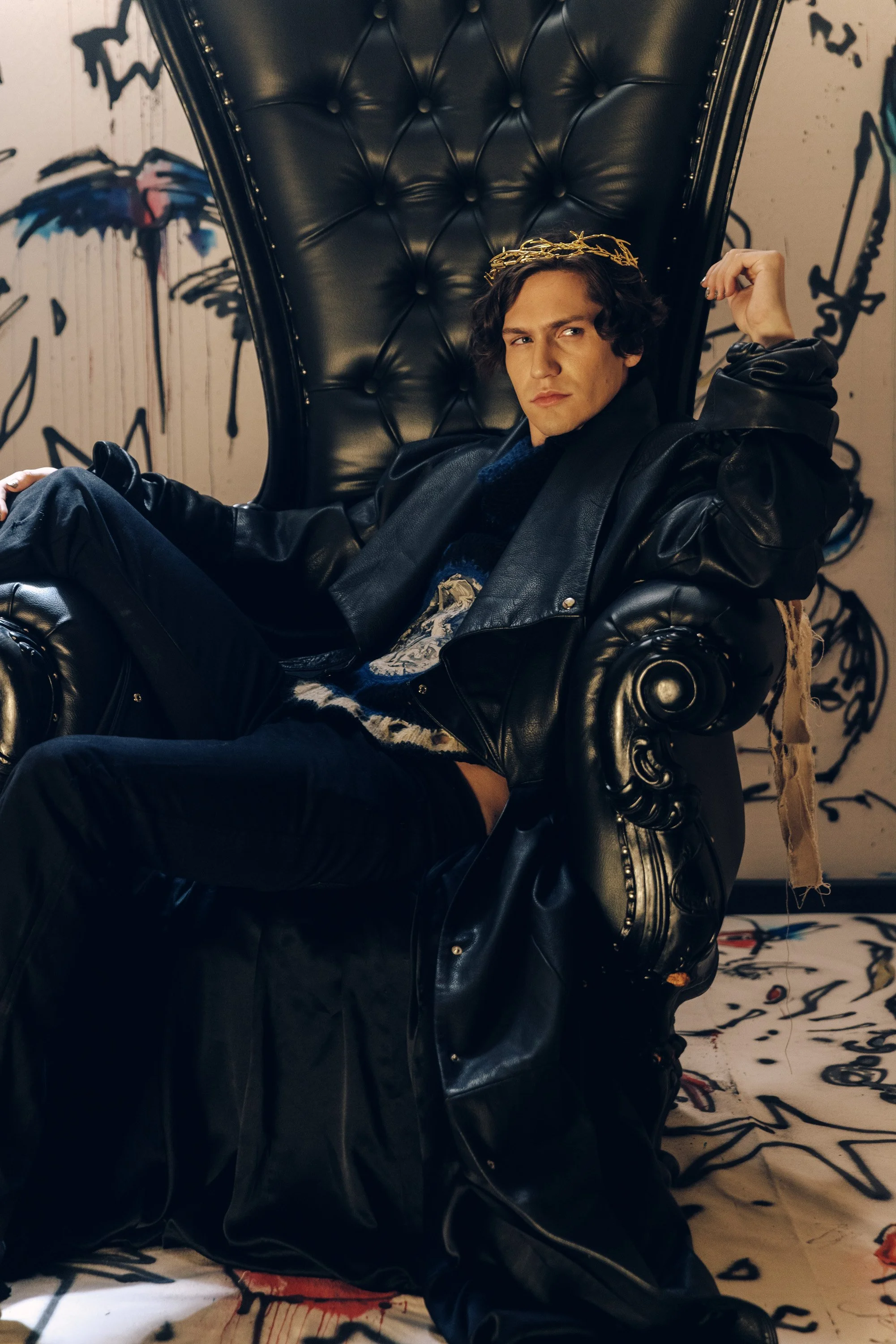 Young man with long curly hair wearing a crown sitting on a large black leather throne with button tufting, in front of a graffiti-style wall.