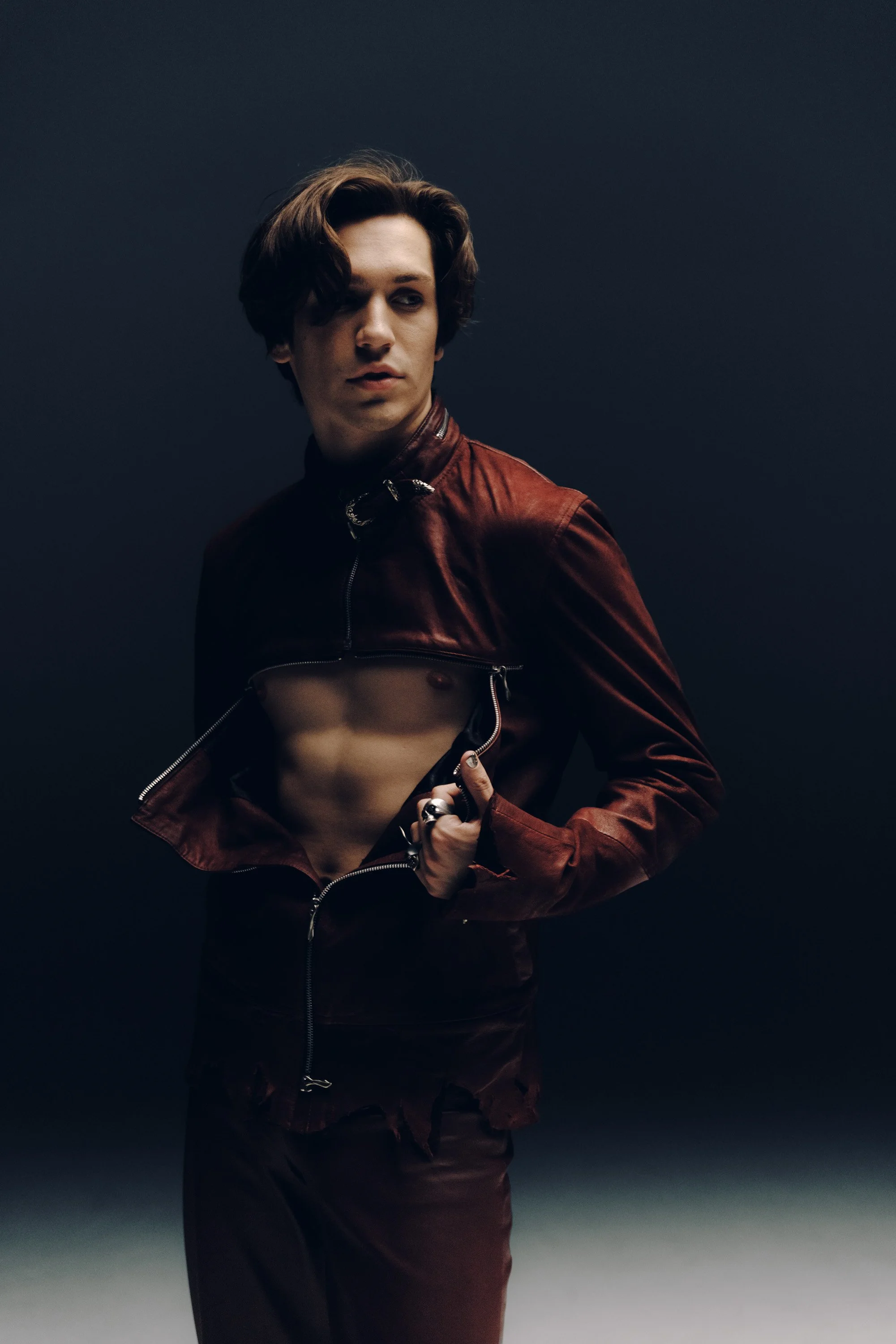 A young man with wavy brown hair and fair skin wearing a partially unzipped brown leather jacket, looking to his right against a dark and gradient background.