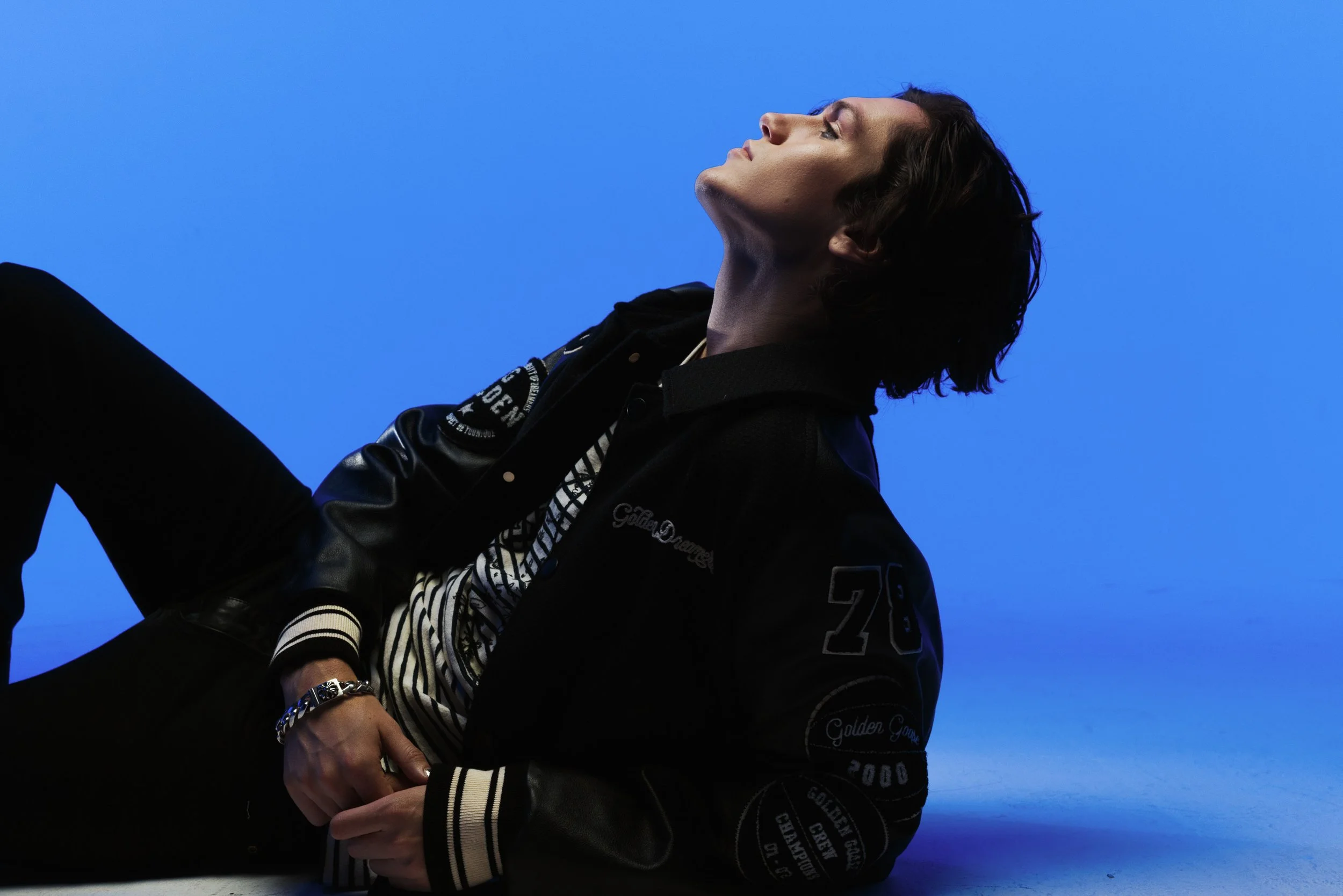 Person with dark, wavy hair wearing a black varsity jacket with patches and a striped shirt, sitting against a bright blue background.