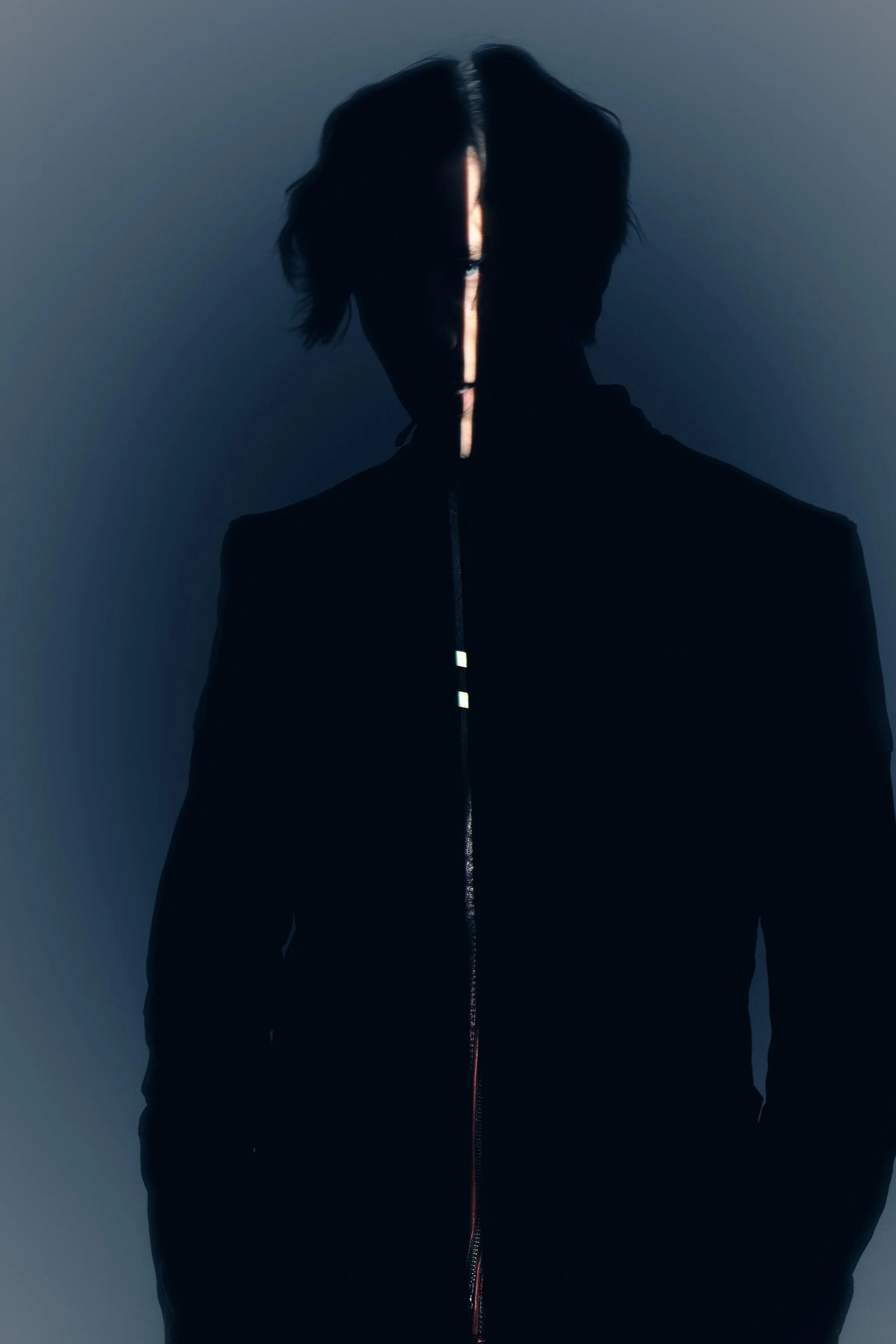 A person wearing a black jacket with a zipper, with their face mostly in shadow and a vertical light streak across their face and chest.