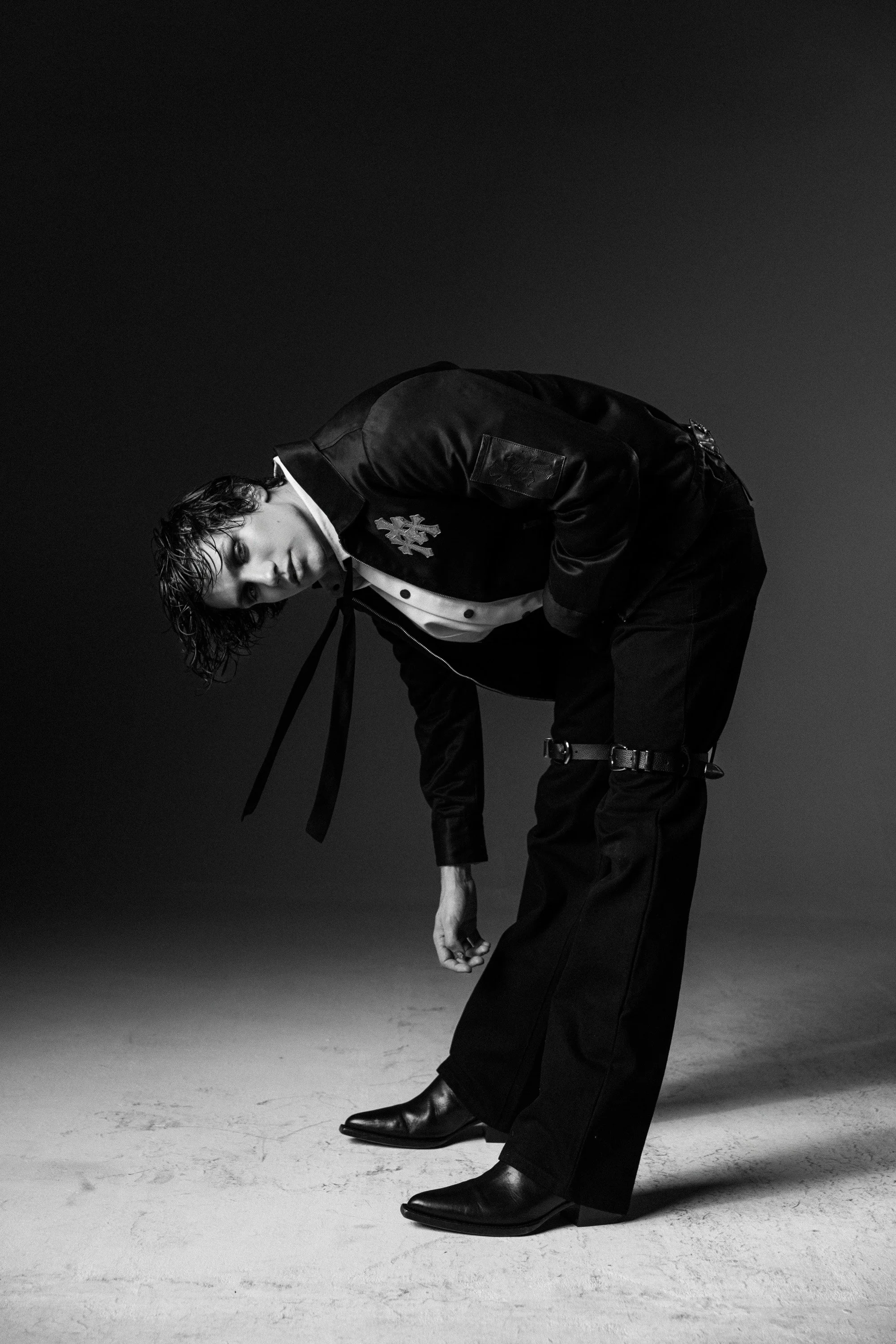 A young person with wet hair bowing over, wearing a satin bomber jacket with a university patch, black pants, and pointed leather boots, against a plain background.