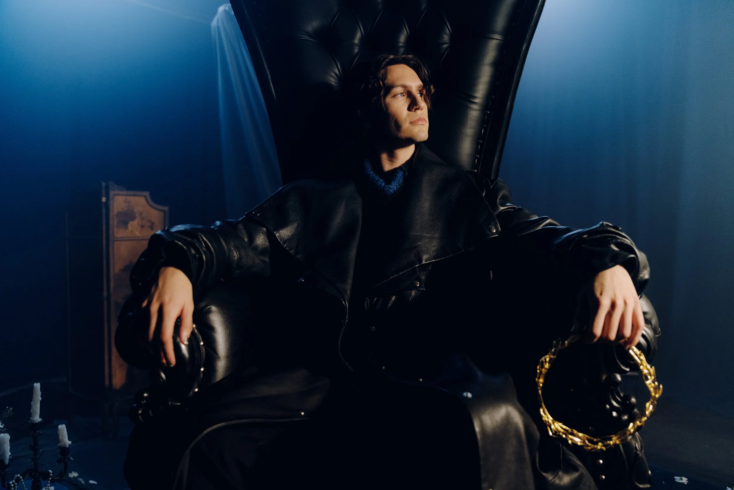 A young man with dark wavy hair sitting on a black leather armchair, wearing a black leather jacket and a dark blue turtleneck. The room has blue-toned lighting and a dark background with a chandelier and a framed picture.