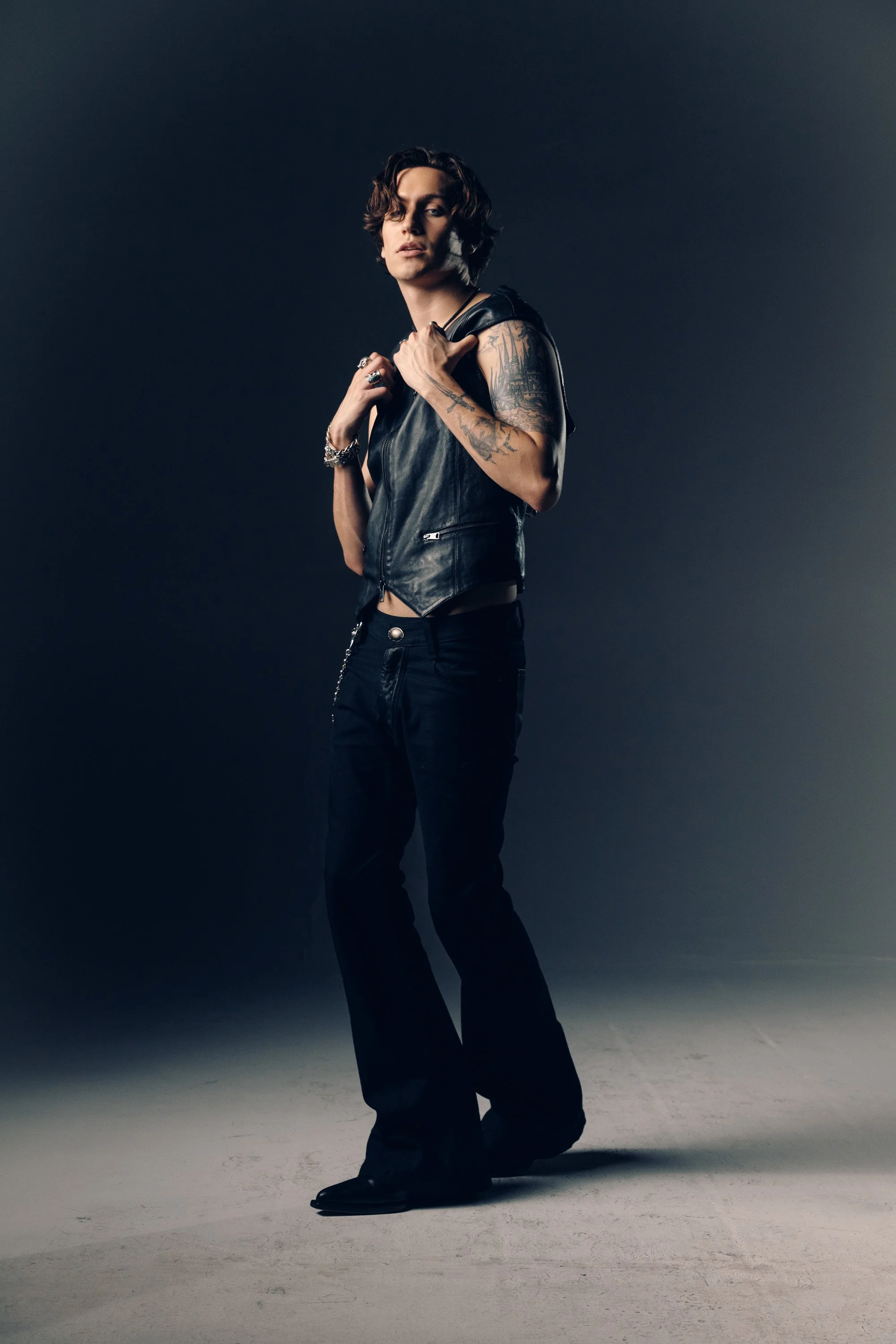 A person with wavy shoulder-length hair, wearing a black leather vest, black pants, and black shoes, standing against a dark background. They have tattoos on their left arm and are accessorized with rings, bracelets, and a chain necklace.
