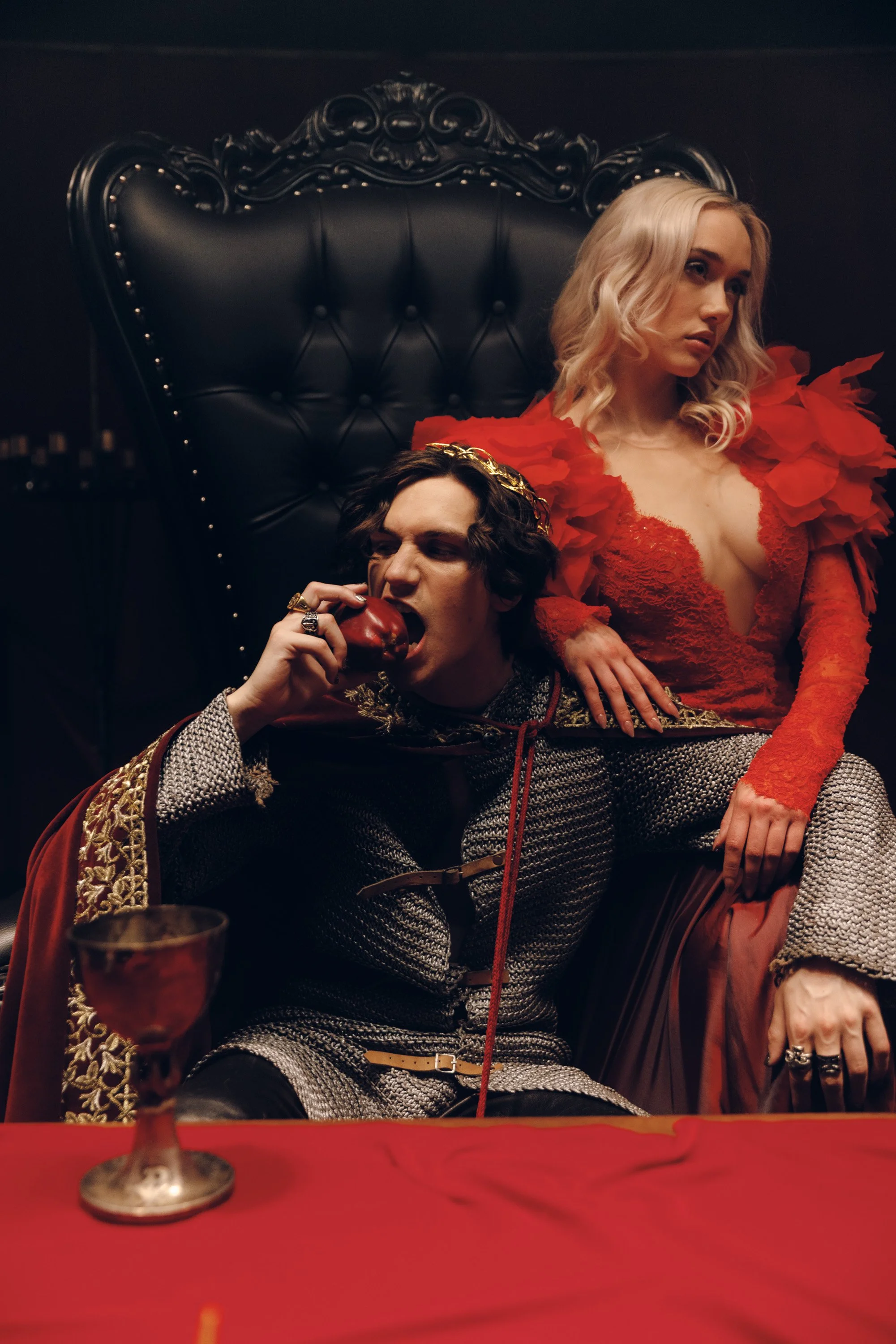 A woman with blonde hair in curls wearing a red dress with ruffled sleeves and a woman with dark hair and gothic attire, sitting at a table with a red tablecloth, an apple, and a glass, in a dimly lit room with a black ornate high-backed chair.