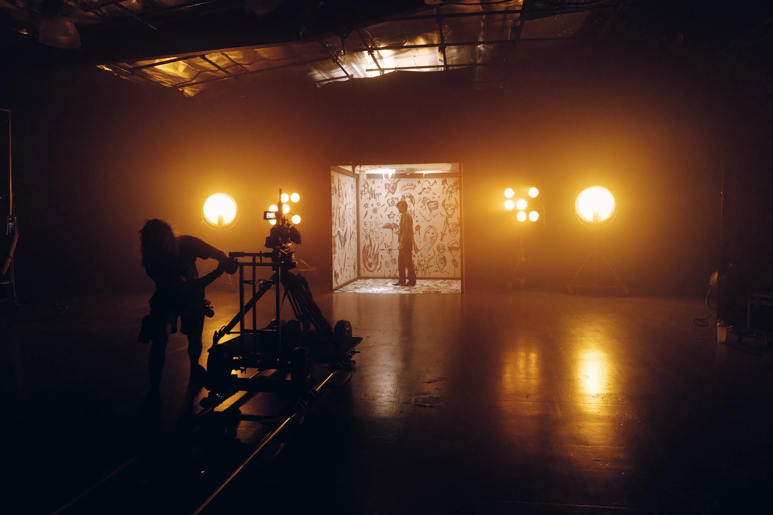 Film production studio with a camera operator, multiple lights, and a backdrop featuring sketches of various characters and objects, illuminated by warm lighting.
