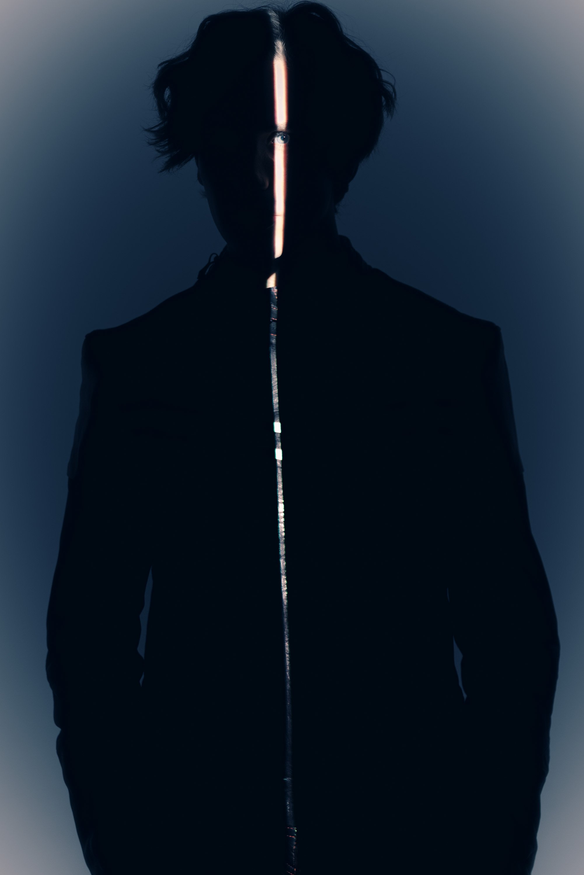 Shadowed person with short hair, wearing a black jacket with a glowing vertical line on the front, in a dimly lit setting.